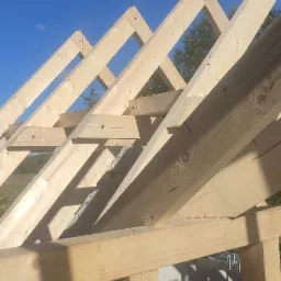 Konstrukcja drewniana więźby dachowej z jasnego drewna, widok od dołu na tle błękitnego nieba i zieleni drzew