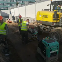 Pracownicy budowlani w kamizelkach odblaskowych używają zagęszczarki wibracyjnej i koparki kołowej Wacker Neuson na placu budowy z widocznymi fundamentami budynku w tle.