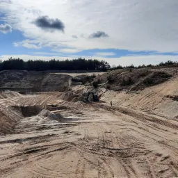 Kopalnia odkrywkowa z jasnym podłożem, ślady opon, koparka w tle i pracownik w jaskrawej kamizelce na tle lasu i błękitnego nieba z chmurami.
