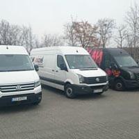 Trzy busy dostawcze zaparkowane obok siebie na placu, jeden biały z widocznym logo firmy, drugi biały, trzeci czarny z grafiką w płomienie, widoczne tablice rejestracyjne.