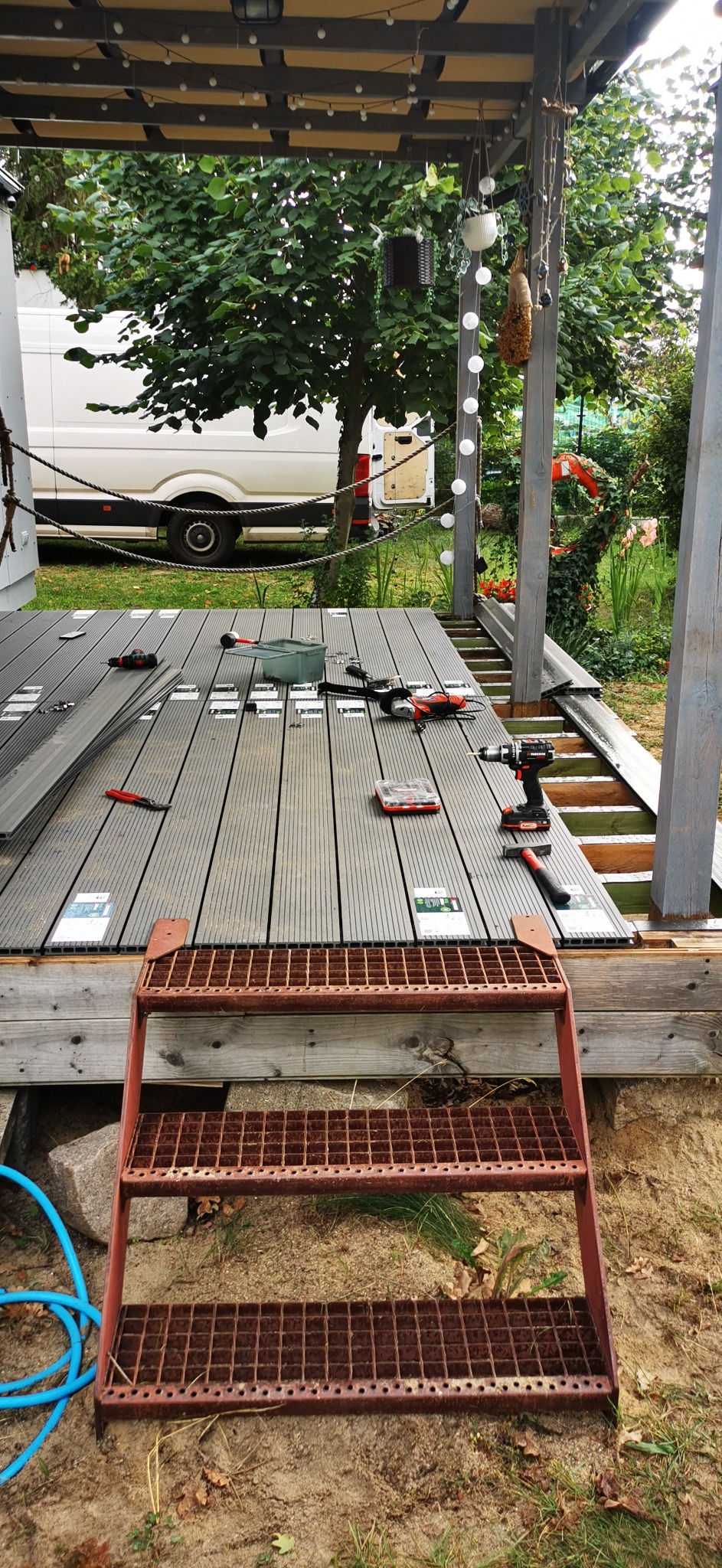 Montaż szarej deski tarasowej na drewnianej konstrukcji, widoczne narzędzia: wkrętarka, młotek, piła, schodki wejściowe, w tle biały bus.