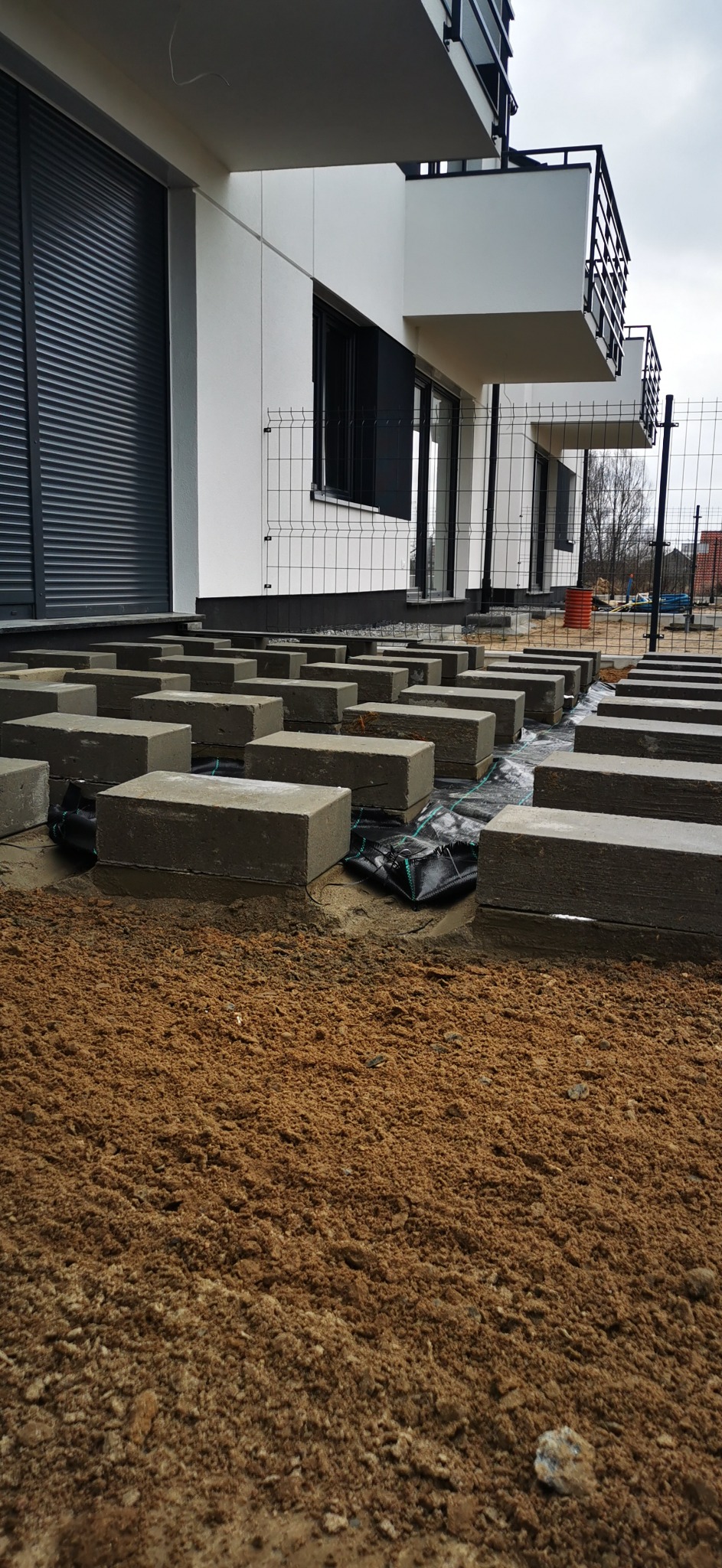 Przygotowanie podłoża pod taras: ułożone betonowe bloczki na geowłókninie, piasek i budynek z balkonami w tle.