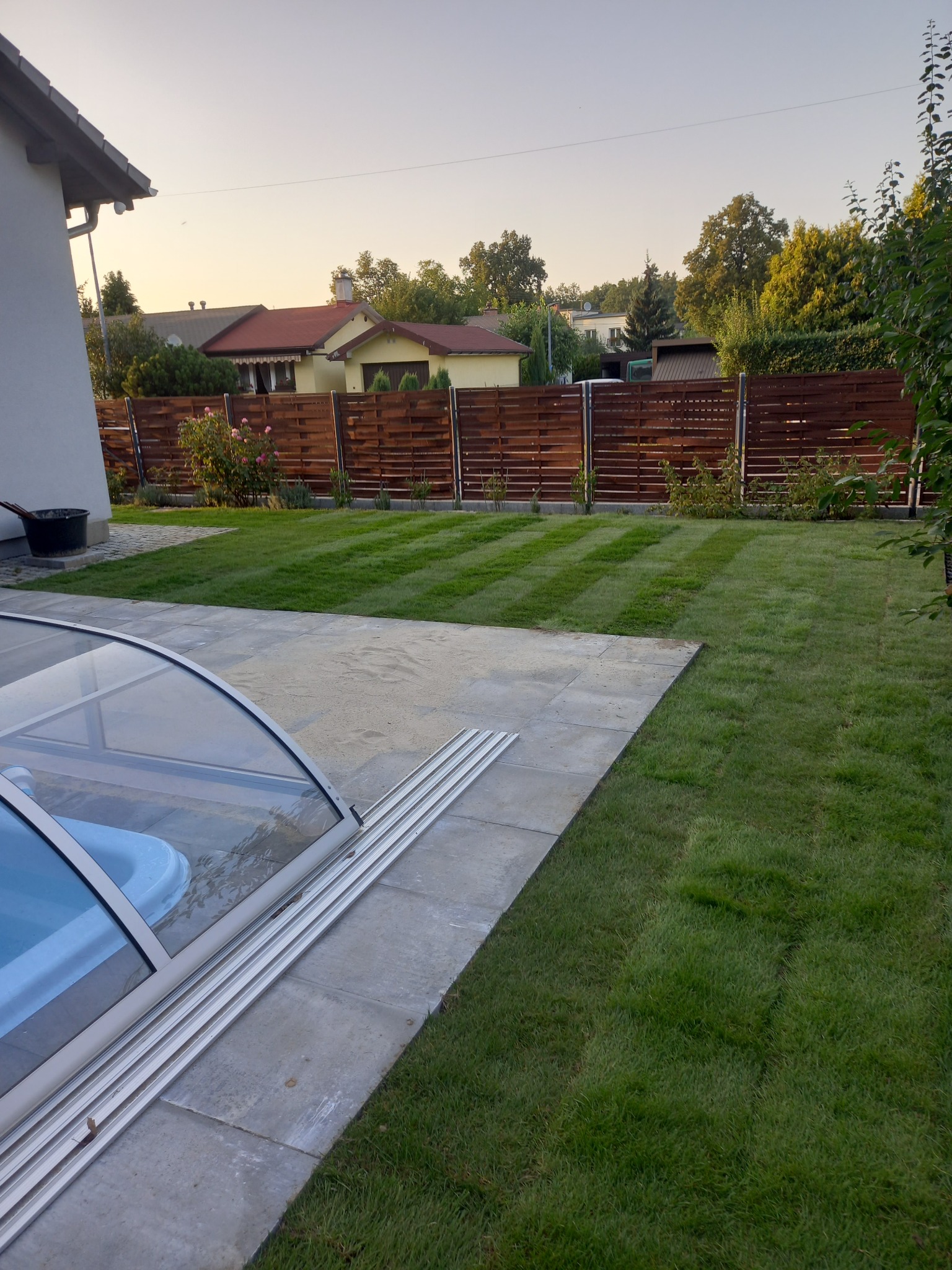 Zadbany ogród z równo przystrzyżonym trawnikiem w pasy, ogrodzony drewnianym płotem, z częściowo otwartą aluminiową konstrukcją zadaszenia basenu.