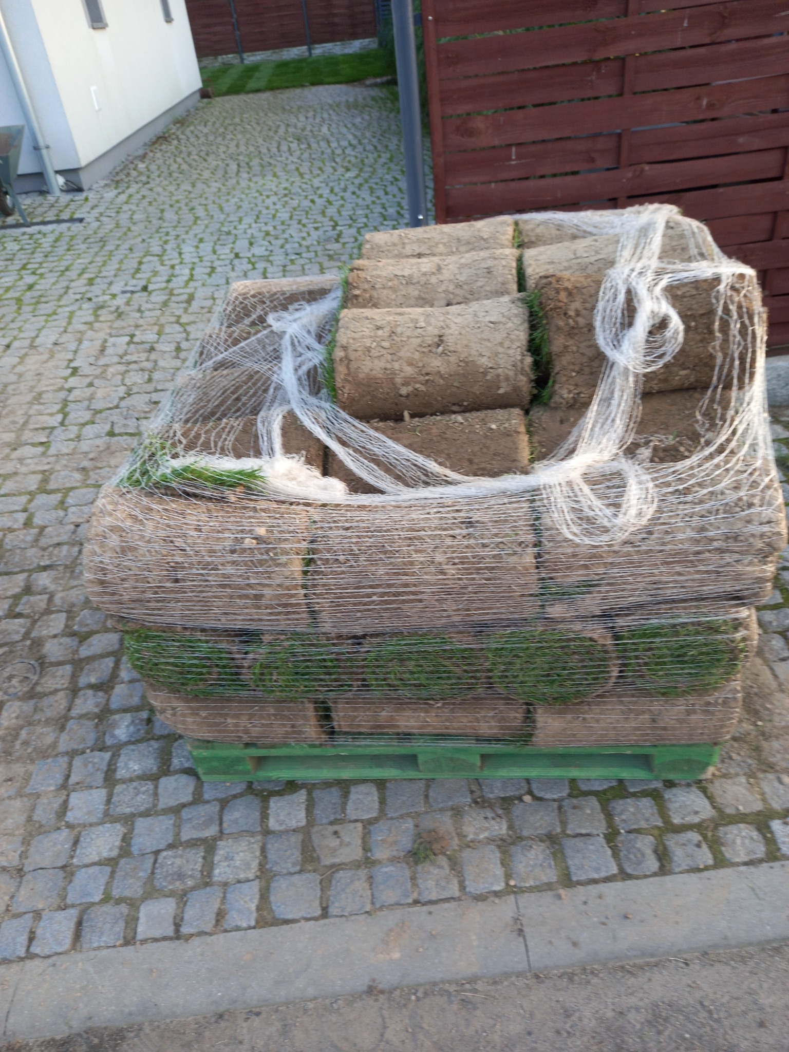 Paleta z rolkami trawy, owinięta folią, leżąca na brukowanej nawierzchni przed domem z drewnianym ogrodzeniem w tle.