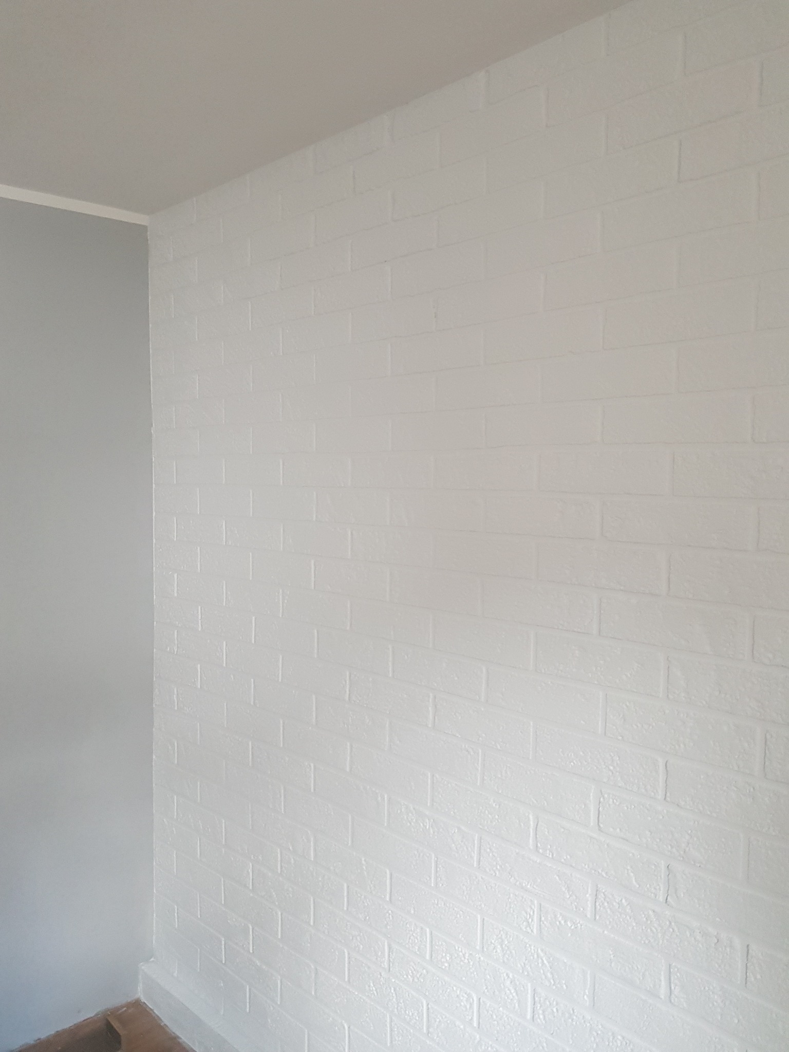 Świeżo pomalowana na biało ściana z efektem cegieł w Radziejowie, z szarą ścianą boczną i listwą przypodłogową, widoczne niedoskonałości faktury.