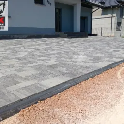 Nowoczesna, szara kostka brukowa ułożona przed domem z numerem 100. Widoczne obrzeża i podsypka. Całość tworzy estetyczny podjazd.