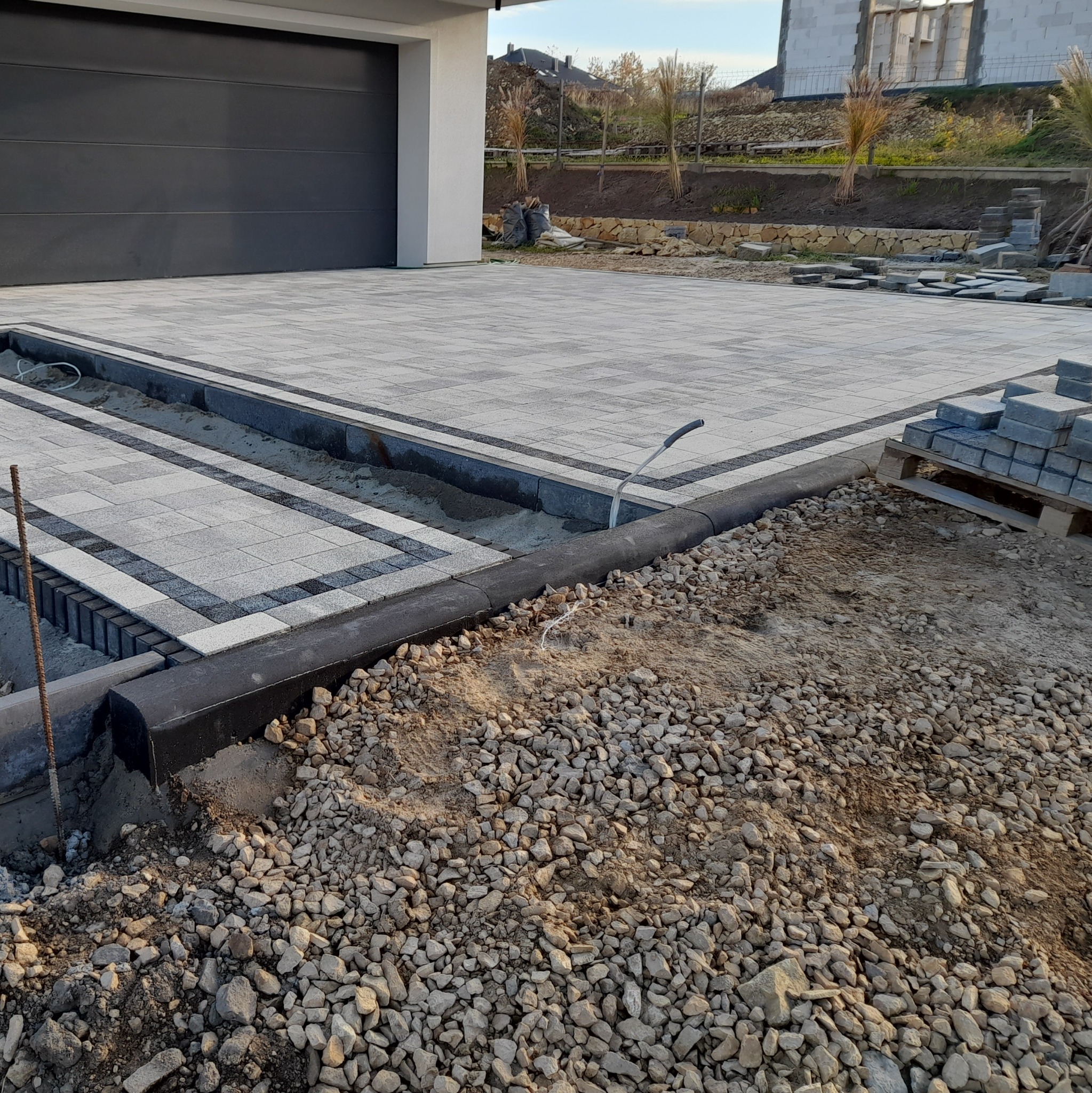 Nowo ułożony podjazd z szarej kostki brukowej z czarną obwódką, w trakcie prac wykończeniowych, widoczne podsypka piaskowa, kamienie i paleta z kostką.