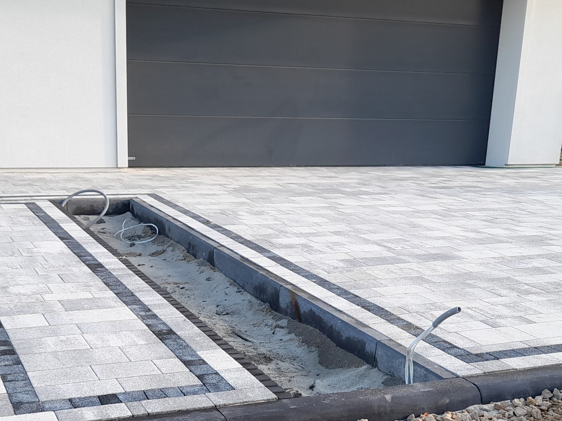 Jasnoszara kostka brukowa ułożona w geometryczny wzór z ciemniejszymi obramowaniami, widoczny fragment kanału odwadniającego wypełnionego piaskiem i wystającymi rurami, brama garażowa w tle.