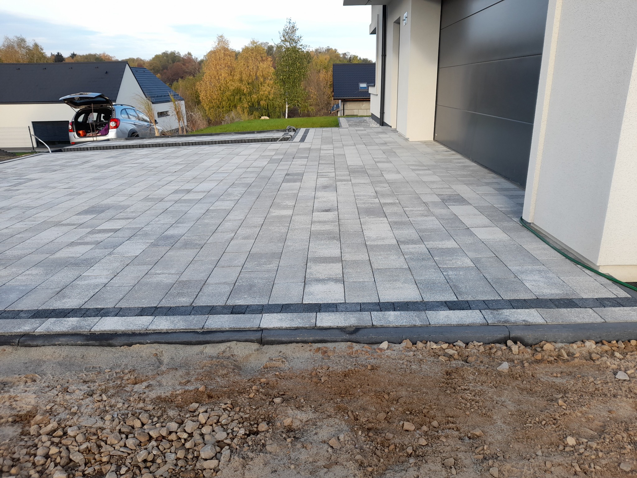 Nowo ułożona, szara kostka brukowa z ciemnoszarym obramowaniem na podjeździe do garażu nowoczesnego domu jednorodzinnego; widoczny samochód z otwartą klapą bagażnika w tle.