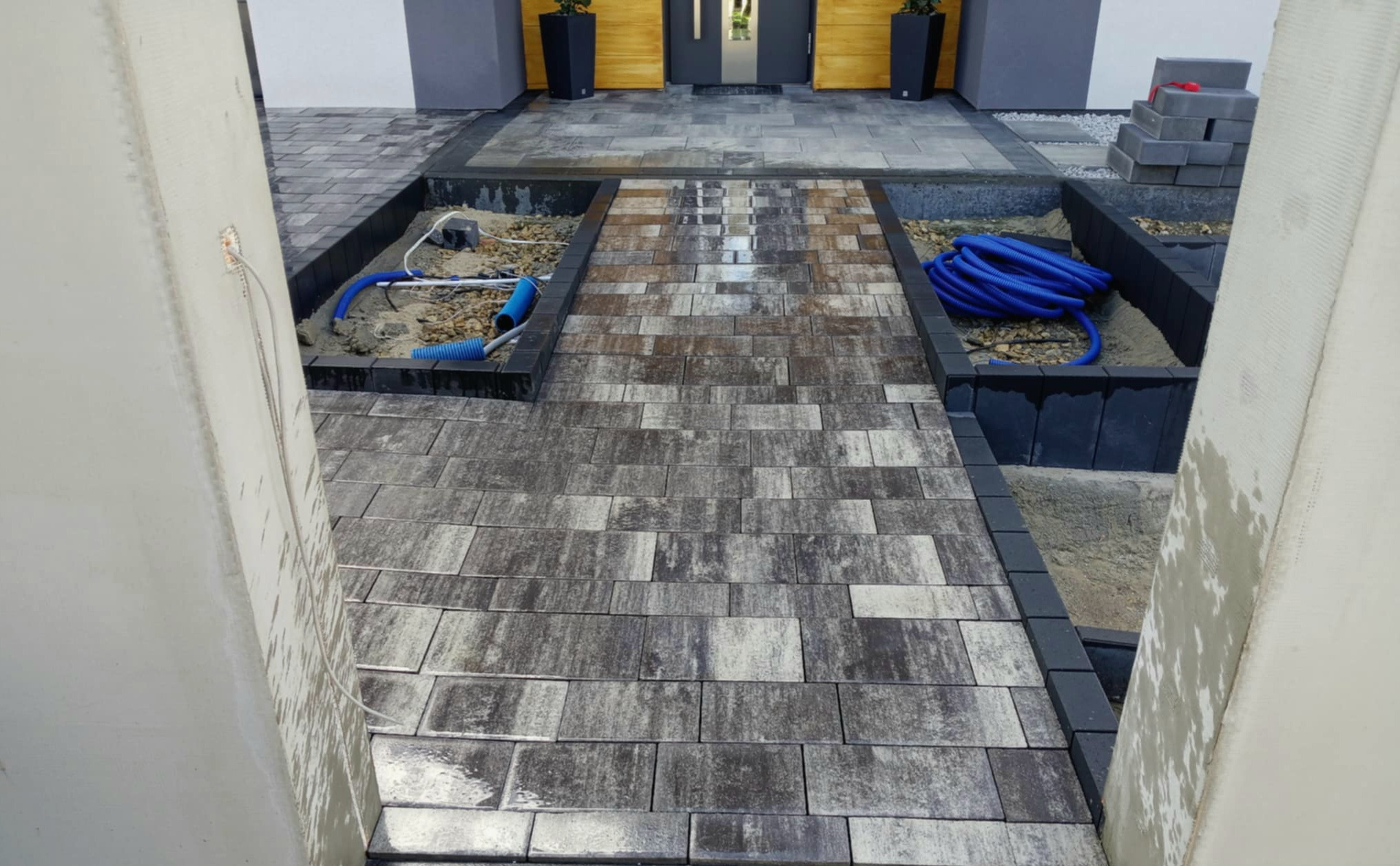 Ułożona kostka brukowa w odcieniach szarości i brązu prowadząca do domu, z widocznymi elementami przygotowania pod instalacje oraz stosem kostki do dalszych prac.