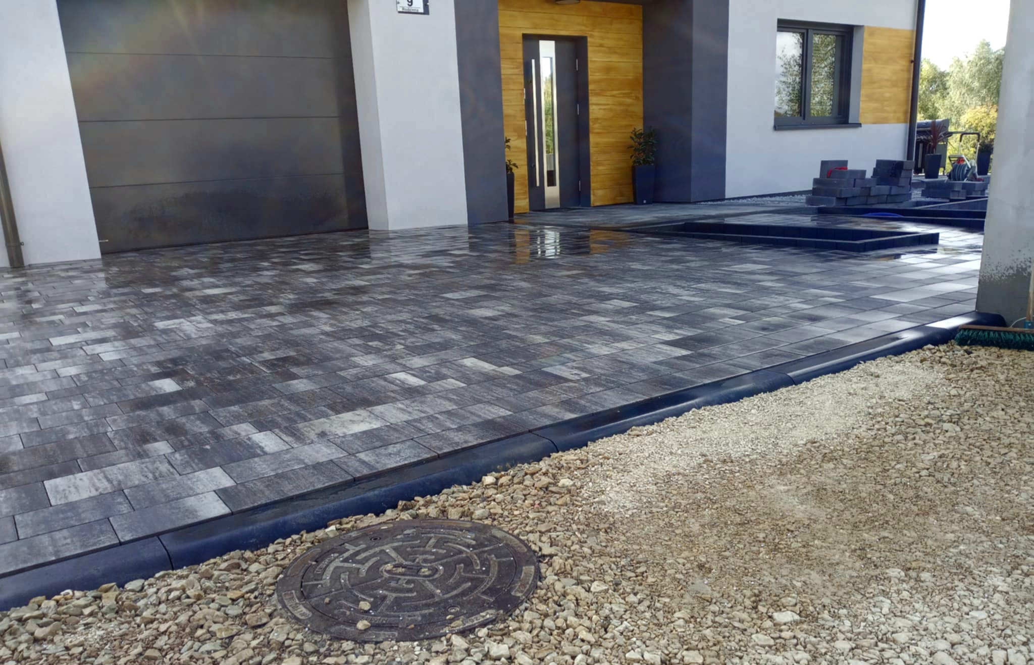 Ułożona szara kostka brukowa na podjeździe przed domem z szarą bramą garażową i drewnianymi drzwiami wejściowymi, obrzeża z czarnego tworzywa, studzienka kanalizacyjna w tle, widoczne kostki...