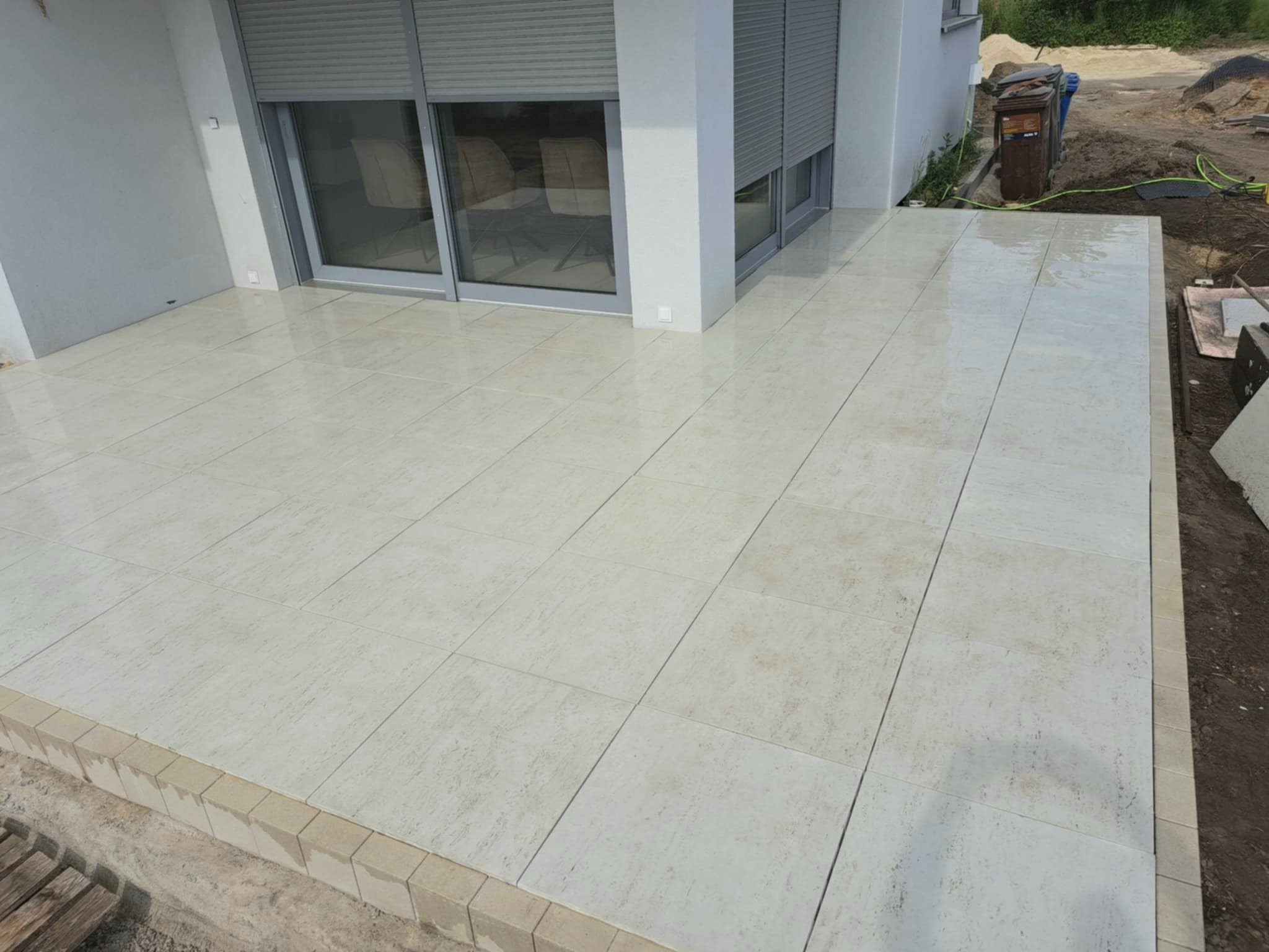 Jasne płytki tarasowe ułożone przy budynku z szarymi roletami, widoczne obrzeże z kostki.