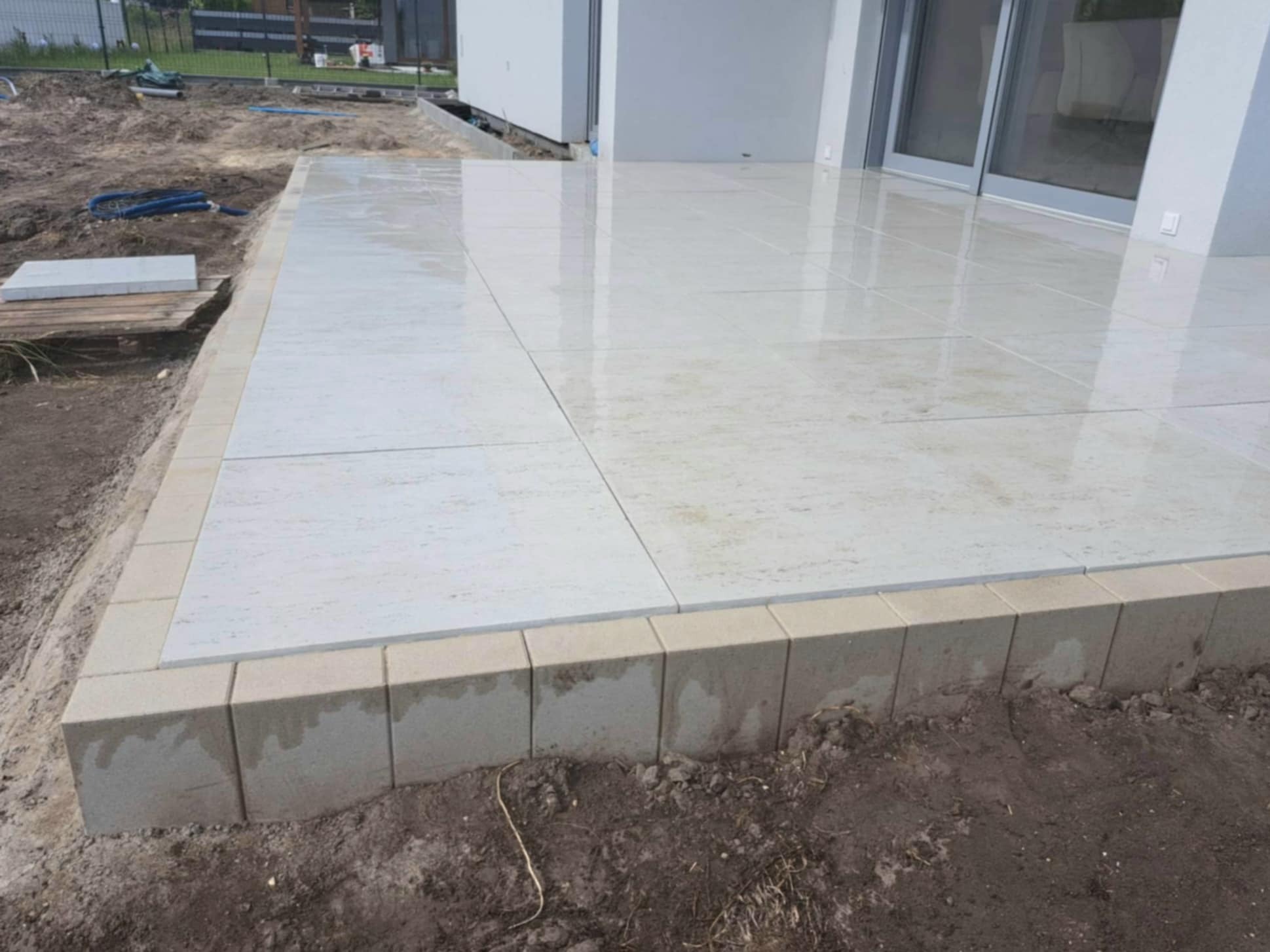 Wyłożony jasnymi płytami taras z obrzeżem z betonowych kostek, widoczne pozostałości ziemi po pracach ziemnych.