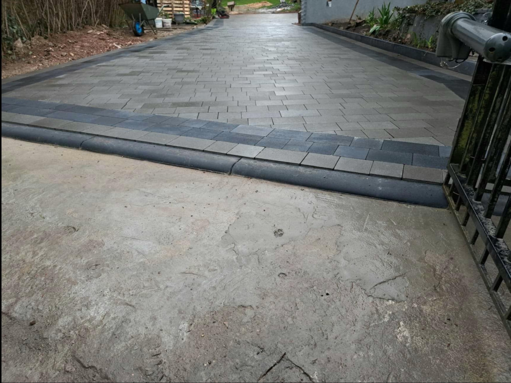 Ułożona nawierzchnia z szarej kostki brukowej wzdłuż podjazdu, z czarnym obrzeżem i widocznym fragmentem betonowej powierzchni przed bramą.