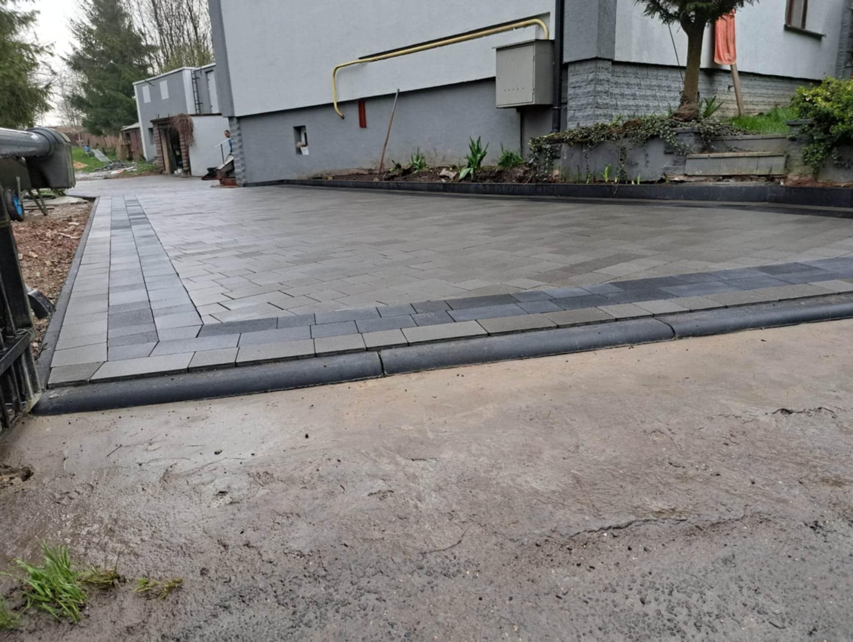 Ułożona szara kostka brukowa przed domem z obrzeżami, widoczny fragment betonowej nawierzchni przed ułożeniem.