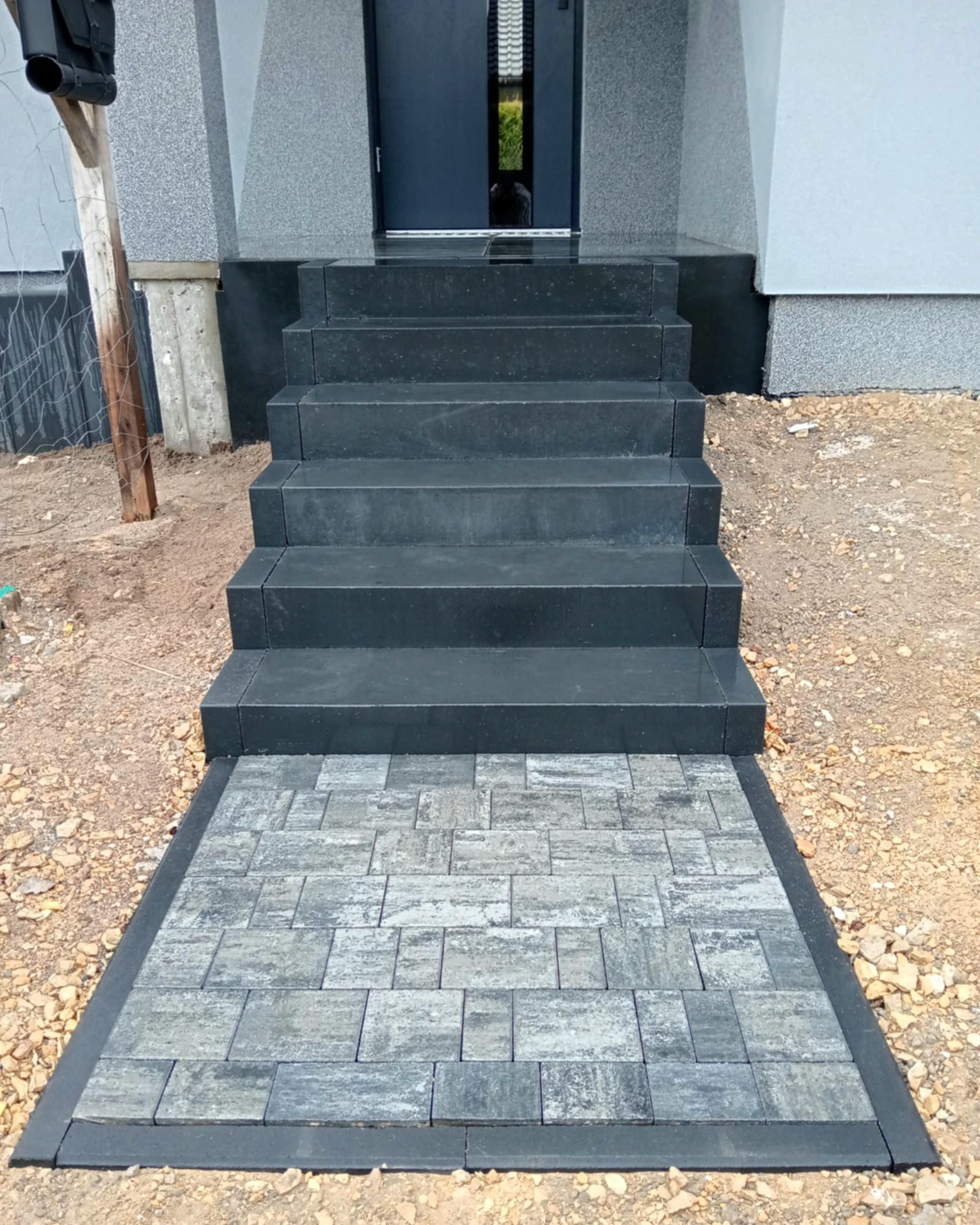 Schody wejściowe z ciemnej kostki prowadzące do drzwi, obrzeża z kostki brukowej w kolorze szarym. Otoczenie w trakcie prac wykończeniowych.