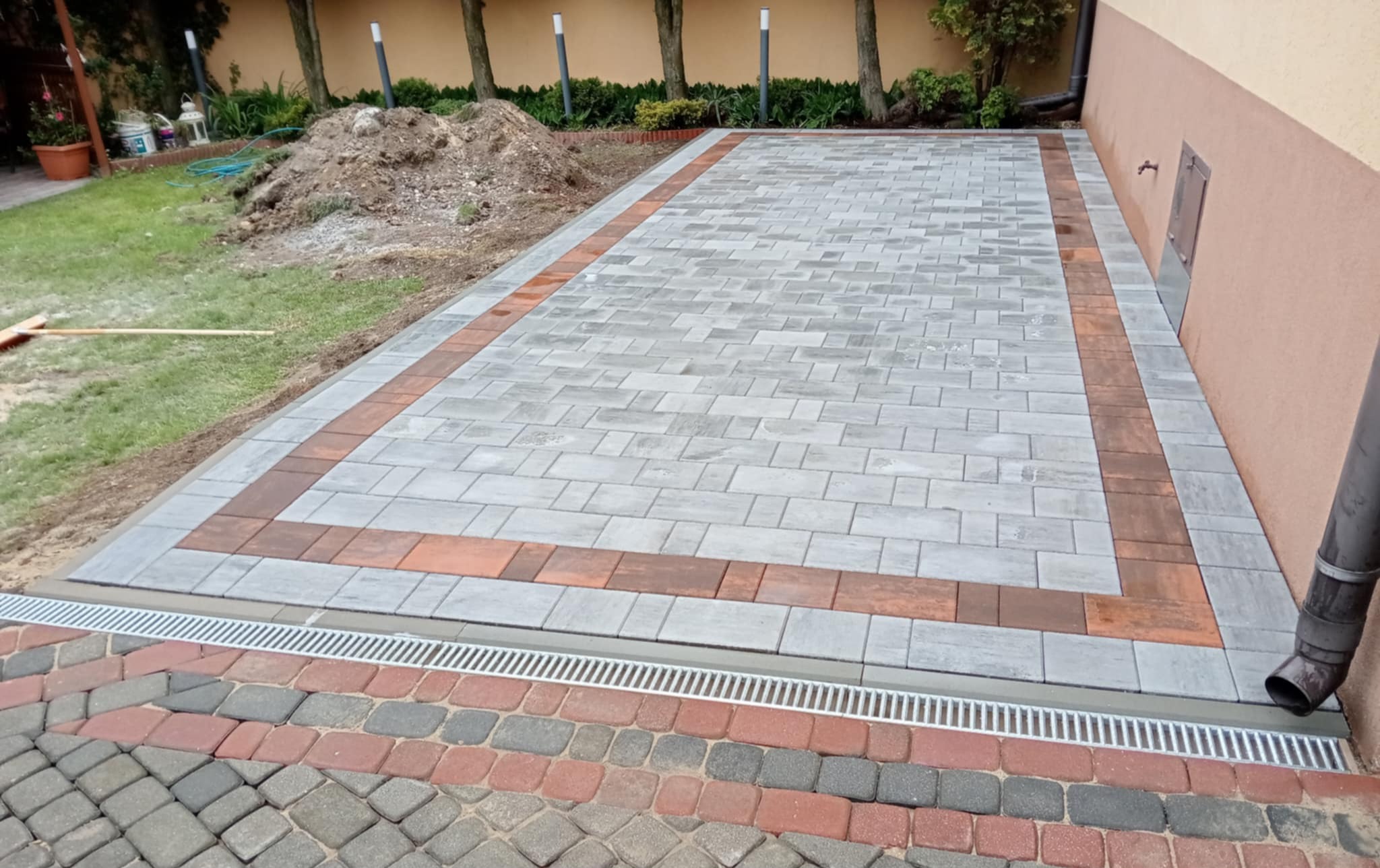 Ułożona kostka brukowa w odcieniach szarości z bordową obwódką, oddzielona od trawnika korytkiem odwadniającym. Częściowo widoczna sterta ziemi i budynek.