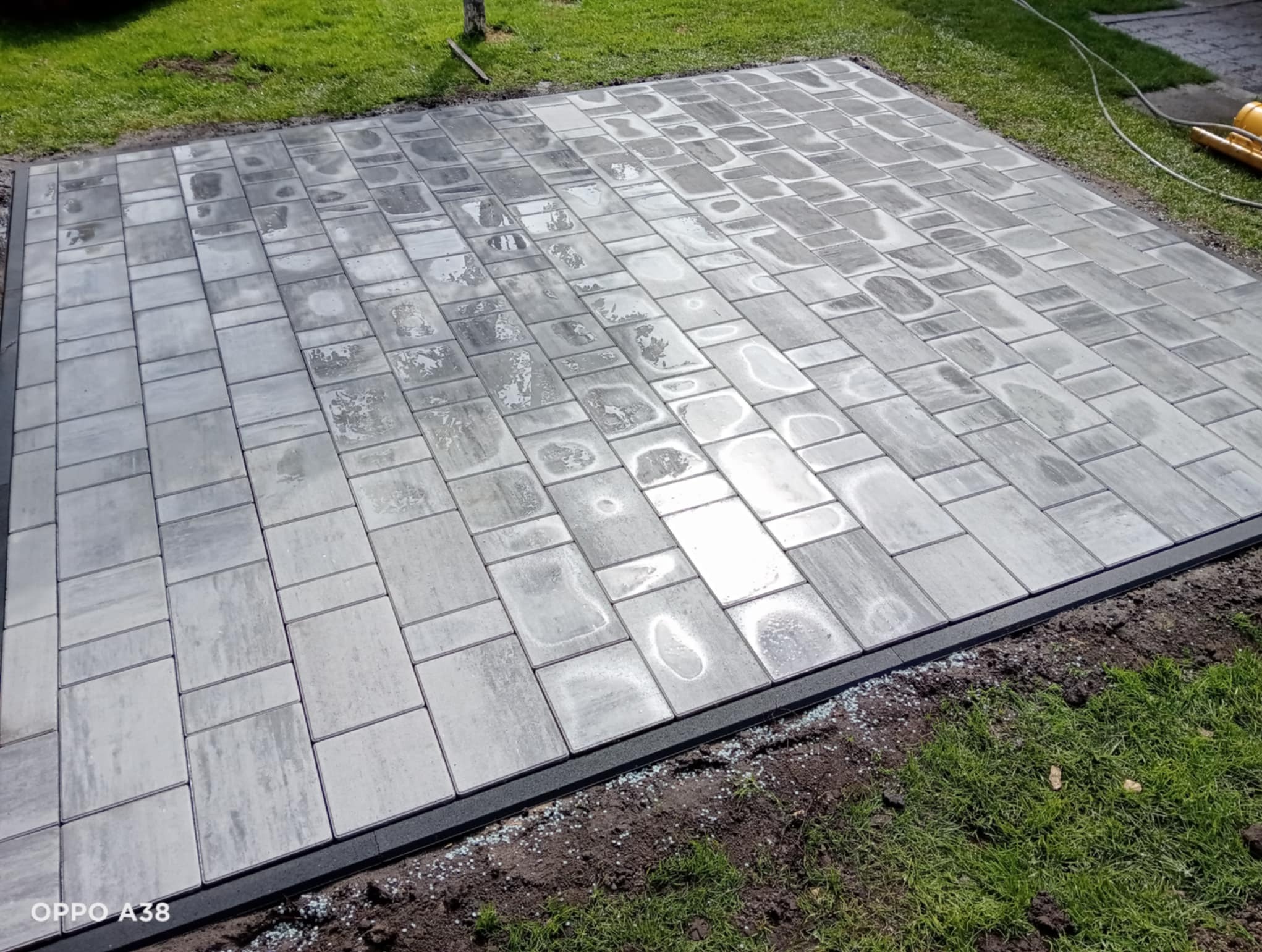 Ułożona szara kostka brukowa na trawniku, widok z góry. Regularny wzór z prostokątnych i kwadratowych elementów. Obrzeże z czarnego tworzywa. Świeżo po deszczu.