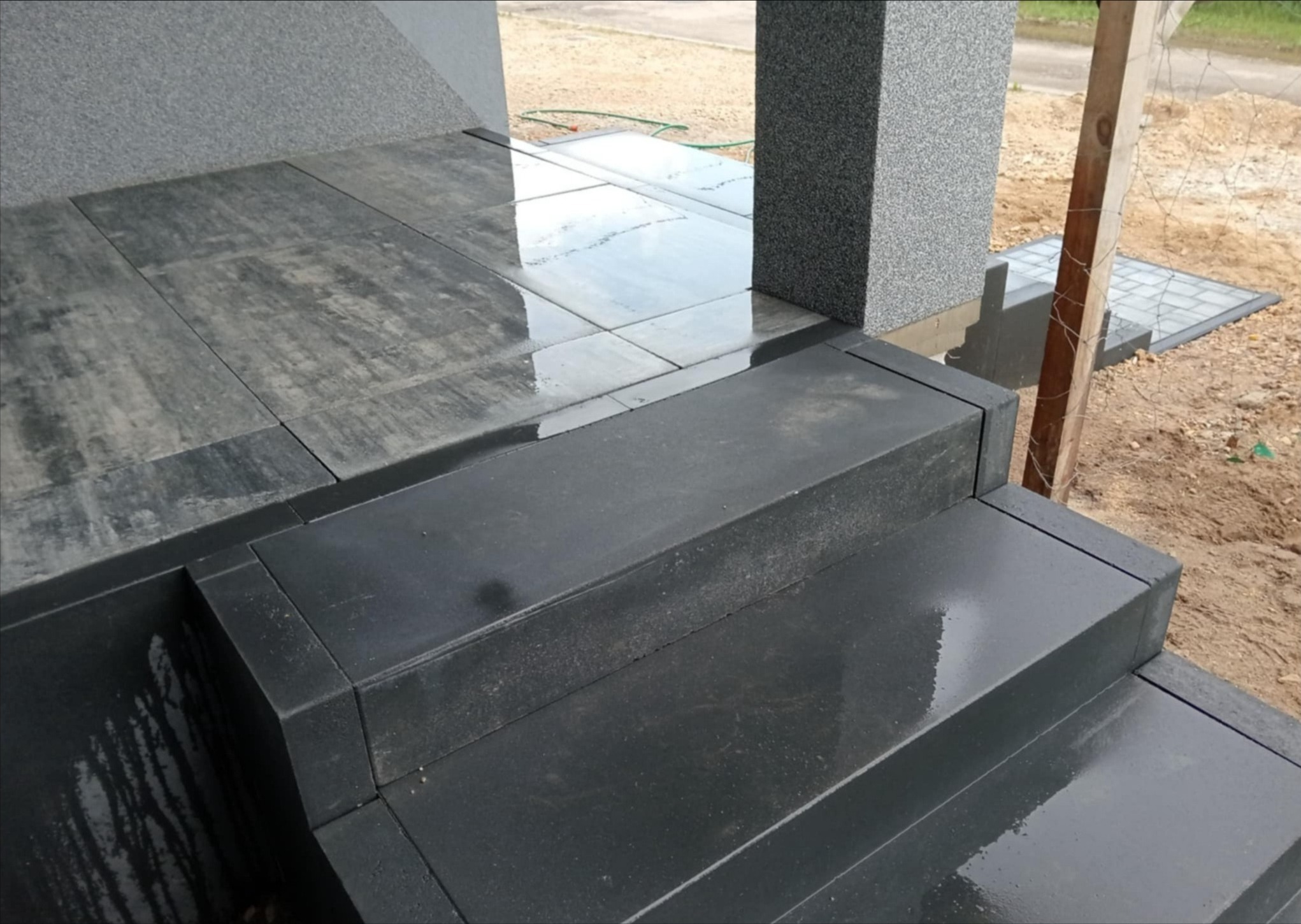 Nowoczesne, grafitowe schody wejściowe z kostki brukowej, z widocznymi refleksami wody po deszczu. Tło stanowi jasna elewacja i fragment terenu budowy.
