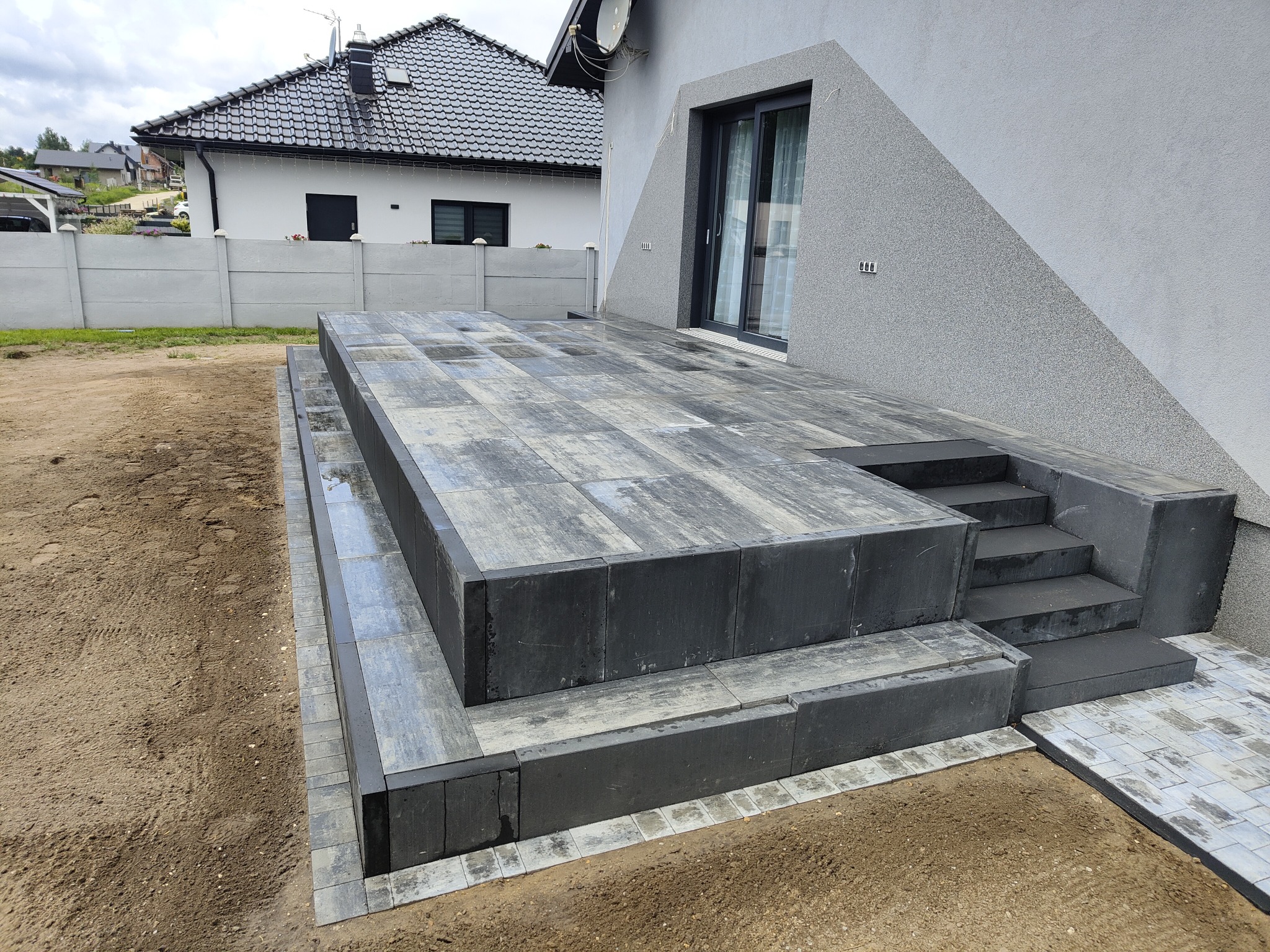 Nowoczesny taras z kostki brukowej w odcieniach szarości, z wyraźnymi schodami i minimalistycznym designem, przylegający do budynku z elewacją w podobnej kolorystyce.
