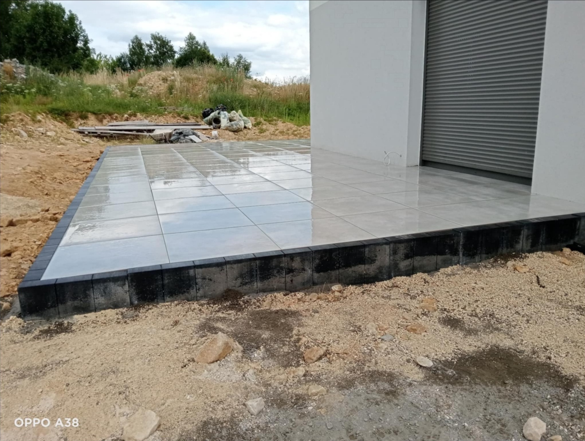 Błyszcząca, jasna kostka ułożona na tarasie z ciemną obwódką. W tle trawa i drzewa. Widoczne niedoskonałości podłoża przed ostatecznym wykończeniem.