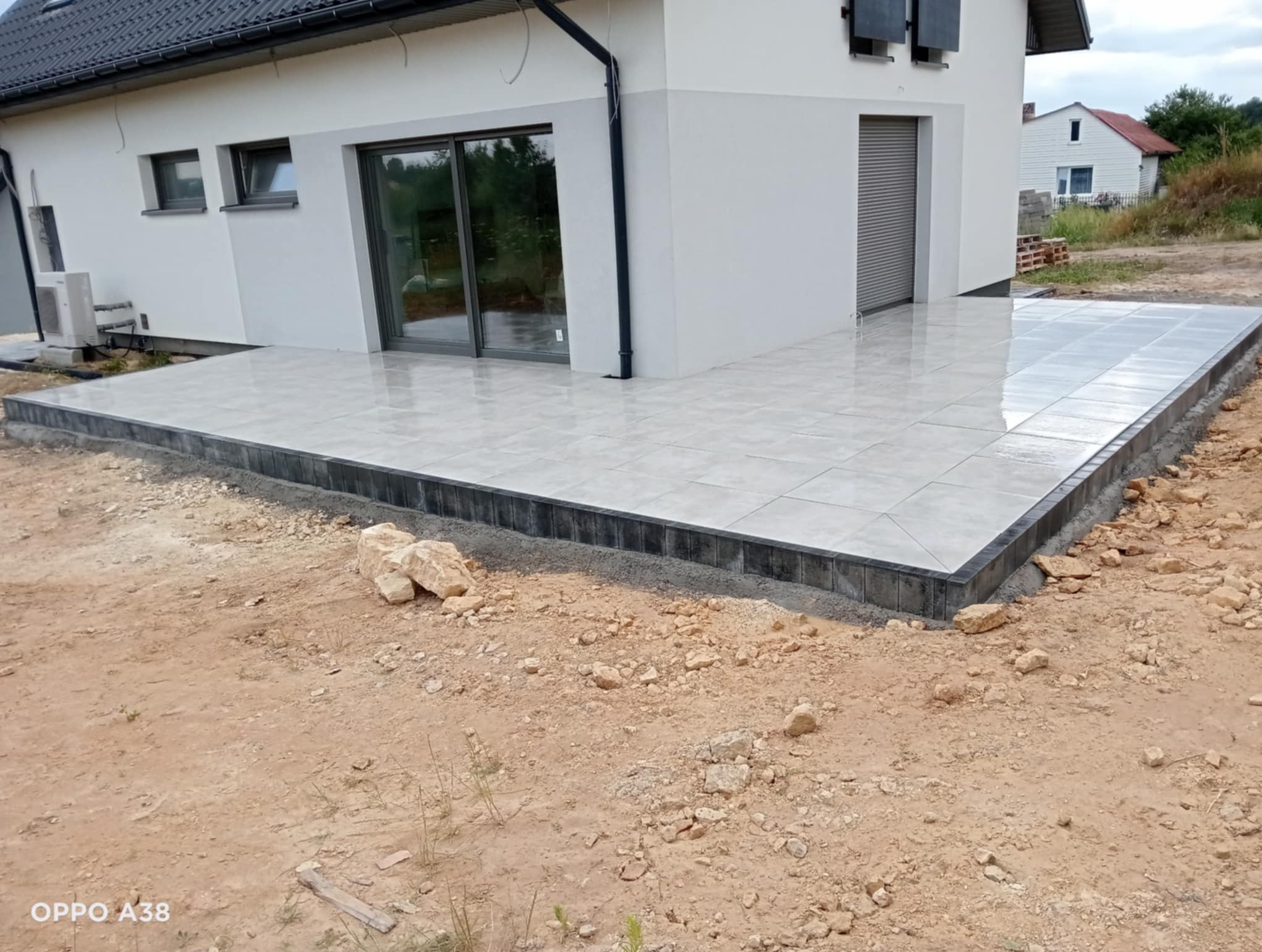 Wykończony taras z jasnych płyt w Strzyżowicach, obrzeża z ciemnej kostki. Widoczne niedokończone otoczenie i fragment domu z oknami.