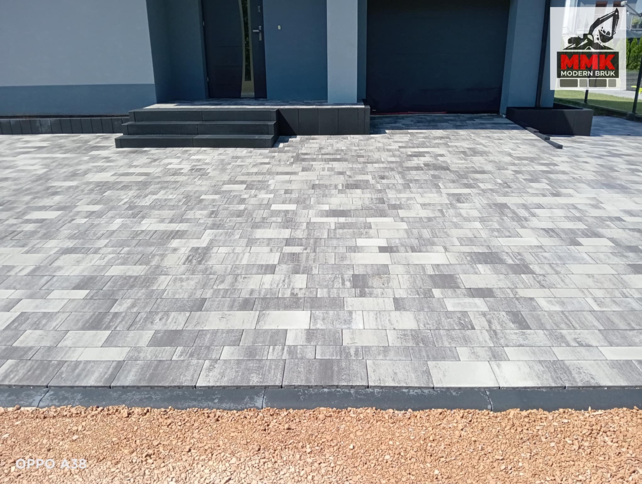 Ułożona szara kostka brukowa z subtelnym wzorem, prowadząca do schodów wejściowych budynku. Widoczny obrzeże i podsypka piaskowa. Całość w nowoczesnym stylu.