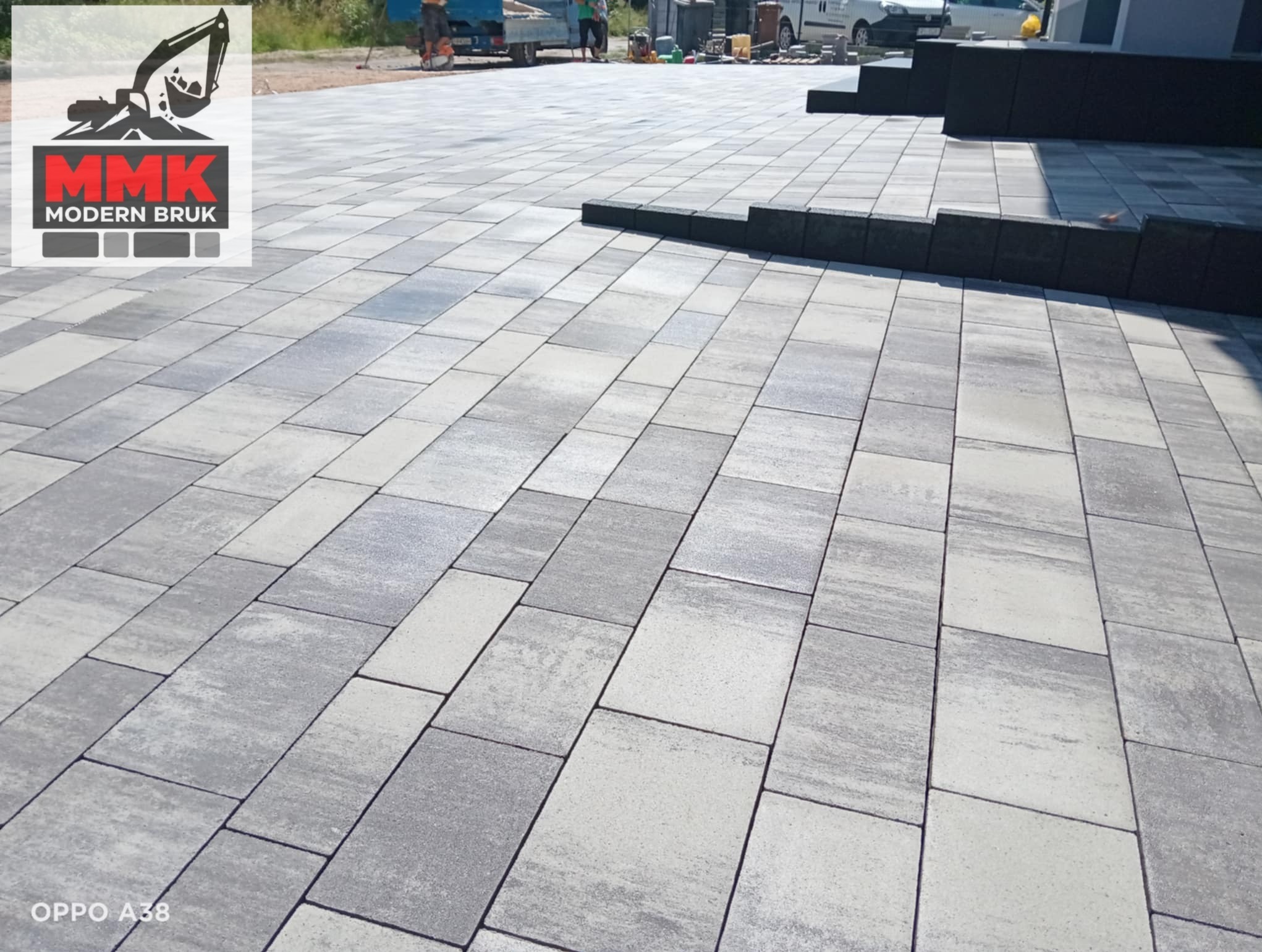 Ułożona, szara kostka brukowa o prostokątnym kształcie, tworząca wzór na podjeździe. W tle widoczne prace brukarskie i sprzęt. Logo firmy MMK Modern Bruk w lewym górnym rogu.