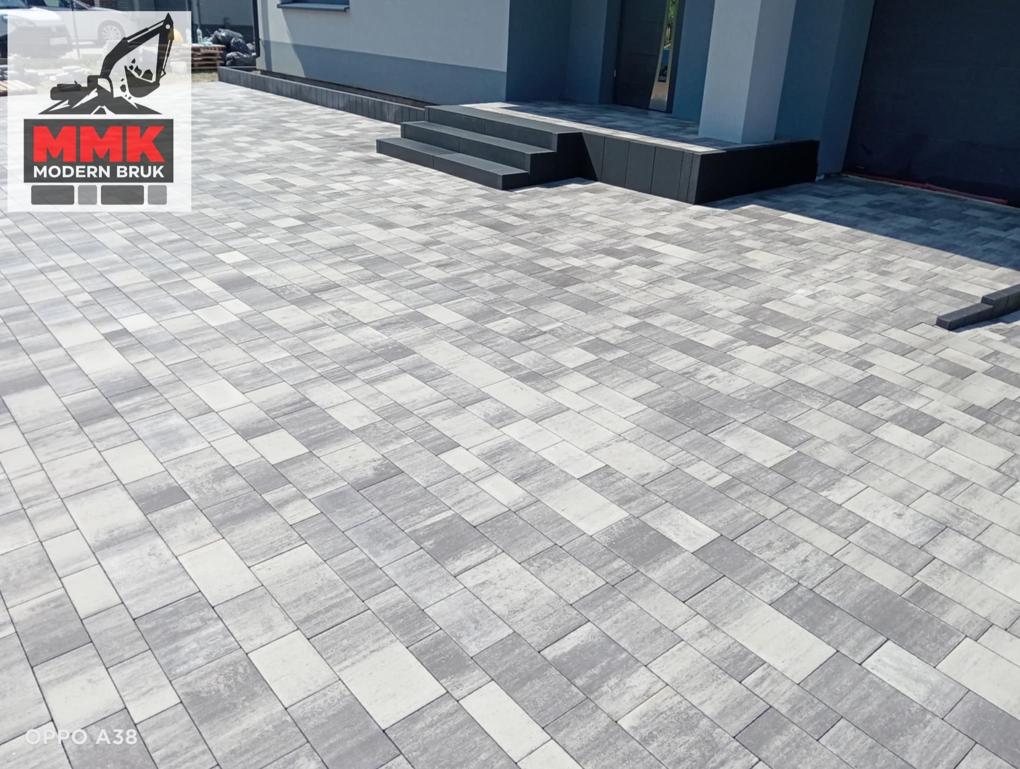 Ułożona, szara kostka brukowa przed nowoczesnym domem z ciemnymi schodami wejściowymi. Widoczny logo firmy MMK Modern Bruk.