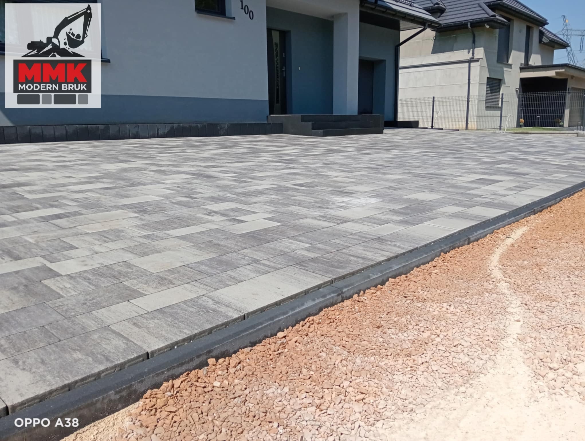 Nowoczesna, szara kostka brukowa ułożona przed domem z numerem 100. Widoczne obrzeża i podsypka. Całość tworzy estetyczny podjazd.