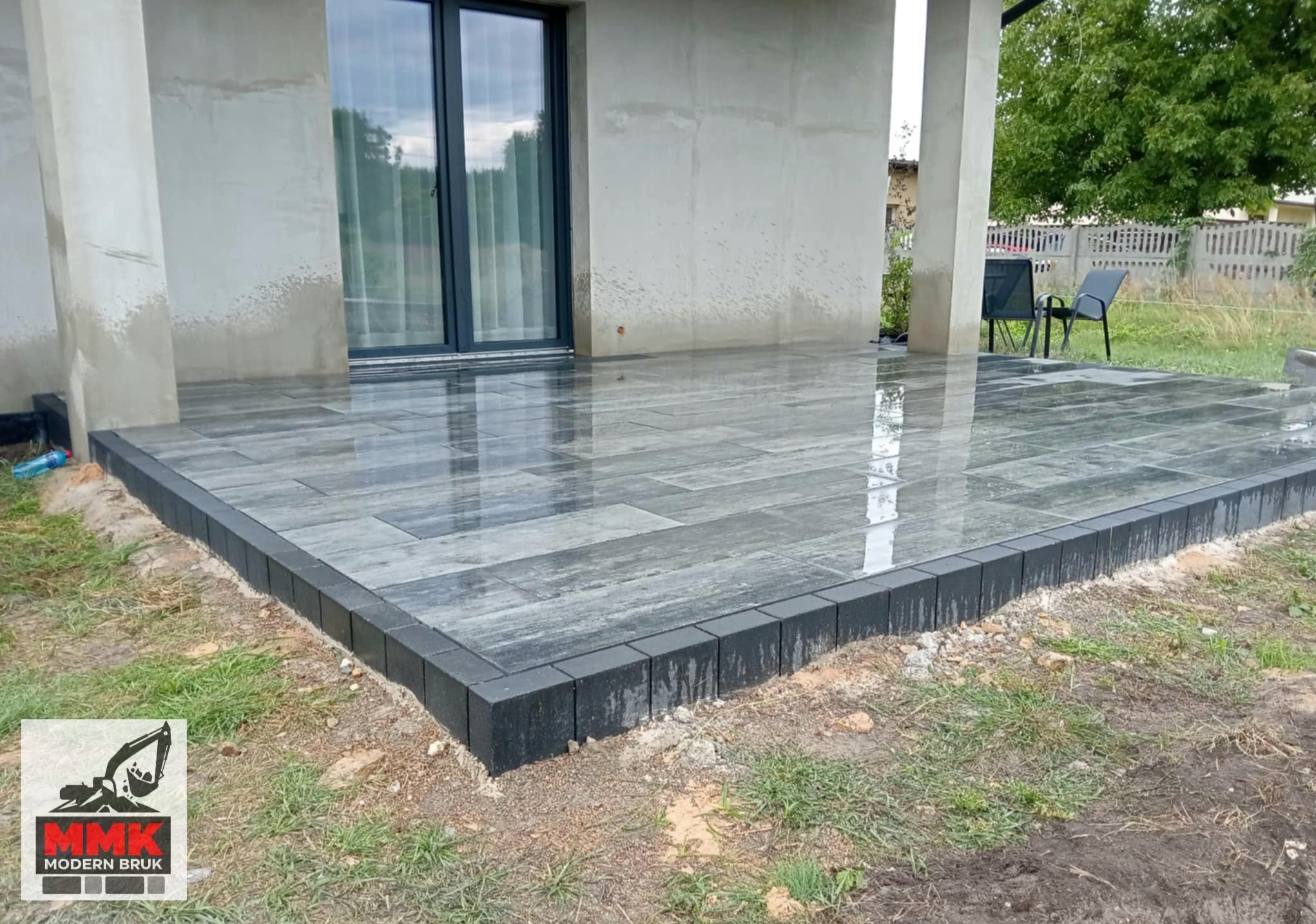 Nowoczesny taras z szarej kostki brukowej ułożonej w prostokątnym wzorze, obrzeża z ciemnej kostki. Widok na dom z dużym oknem tarasowym i zielony ogród.