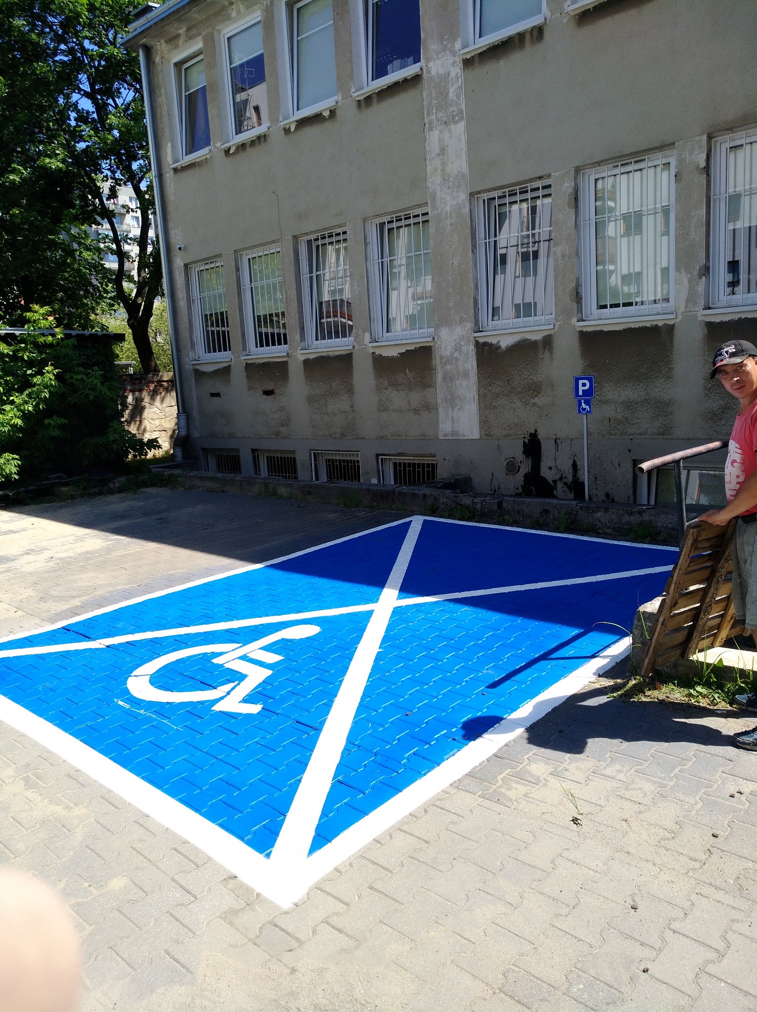Oznakowane na niebiesko miejsce parkingowe dla osoby niepełnosprawnej, biały piktogram i obramowanie, widoczny pracownik z drabinką, budynek w tle.