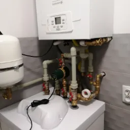 Kompaktowy, biały piec gazowy De Dietrich zawieszony na ścianie z szarymi płytkami, z widoczną instalacją hydrauliczną z mosiężnymi zaworami, rurami z tworzywa i pompą obiegową DAB Evosta 2,...