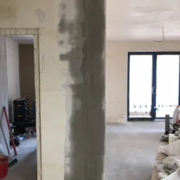 Wnętrze w trakcie prac przygotowawczych: częściowo nałożona szara masa szpachlowa na ścianie i filarze, okno zabezpieczone folią i taśmą, widoczne narzędzia i materiały budowlane.