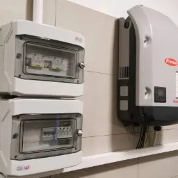 Dwa białe rozdzielnice elektryczne ETI z widocznymi bezpiecznikami, zamontowane jeden nad drugim na kafelkowanej ścianie, obok szary inwerter Fronius SYMO z podłączonymi kablami.