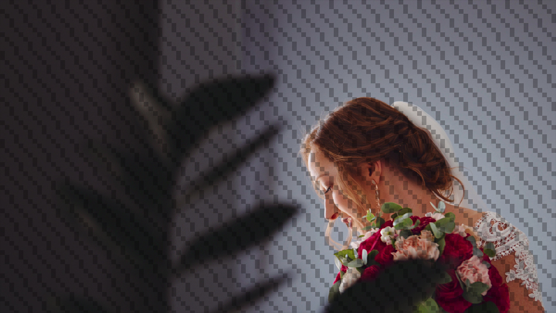 Panna młoda z rudymi włosami w białej sukni z koronkowymi rękawami, trzyma bukiet czerwonych i różowych róż, pochylona głowa, zamyślony profil na tle szarej ściany z geometrycznym wzorem.