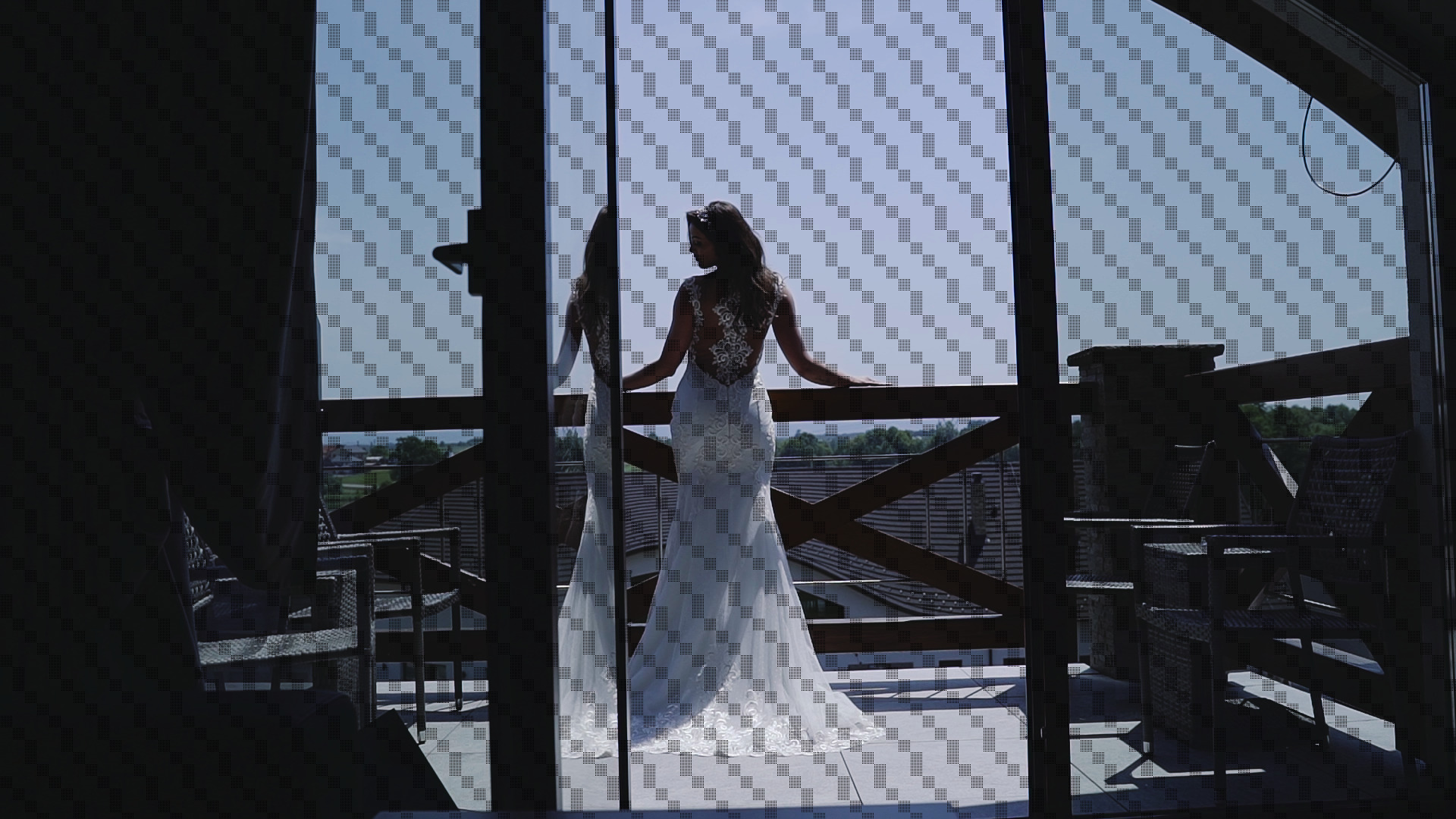 Panna młoda w koronkowej sukni, stojąca na balkonie z drewnianą balustradą, widoczna przez szklane drzwi z refleksami, w tle krajobraz.