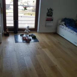 Pokój dziecięcy z drewnianą podłogą, białymi meblami, łóżkiem z dinozaurami i rozłożonymi klockami Lego na czarnej macie do zabawy.