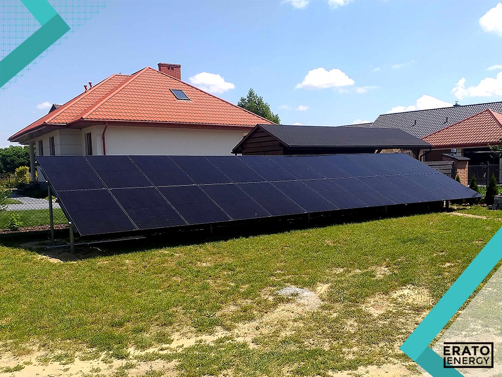 Instalacja paneli słonecznych na gruncie, widok z boku, na tle domów jednorodzinnych w słoneczny dzień.