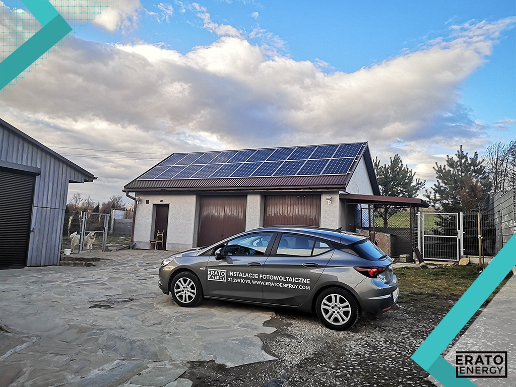 Szary samochód z logo firmy instalującej panele słoneczne zaparkowany przed budynkiem z zamontowaną instalacją fotowoltaiczną na dachu, widoczne dwa psy za ogrodzeniem.