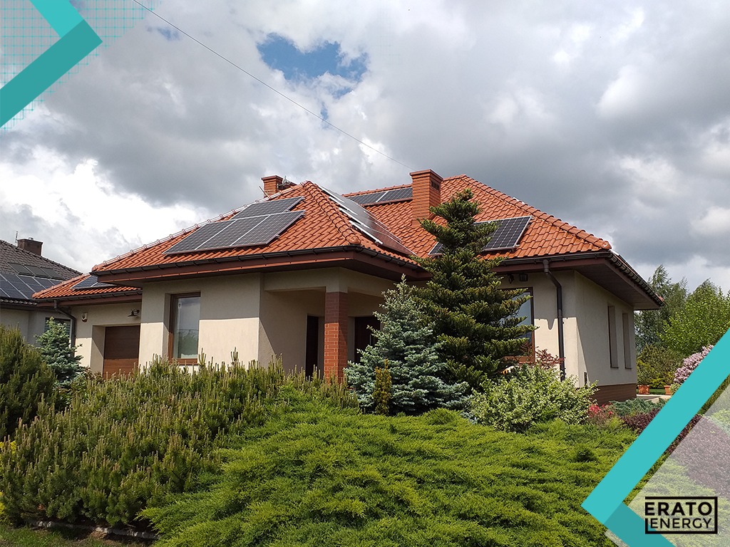 Dom jednorodzinny z czerwonym dachem pokrytym panelami fotowoltaicznymi, widoczny z poziomu ogrodu z bujną zielenią i ozdobnymi iglakami.