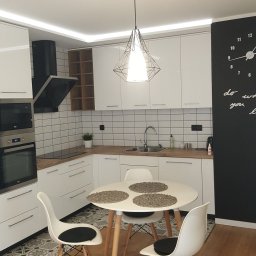Perfect Sprzątanie profesjonalne - Jasna kuchnia z białymi meblami, okrągłym stołem z trzema plecionymi podkładkami i trzema krzesłami z czarnymi poduszkami, czarny zegar ścienny z napisem.