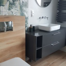 Perfect Sprzątanie profesjonalne - Nowoczesna łazienka z drewnopodobnymi płytkami podłogowymi i ściennymi, umywalką nablatową na szarej szafce z szufladami, okrągłym lustrem z podświetleniem LED oraz toaletą podwieszaną.