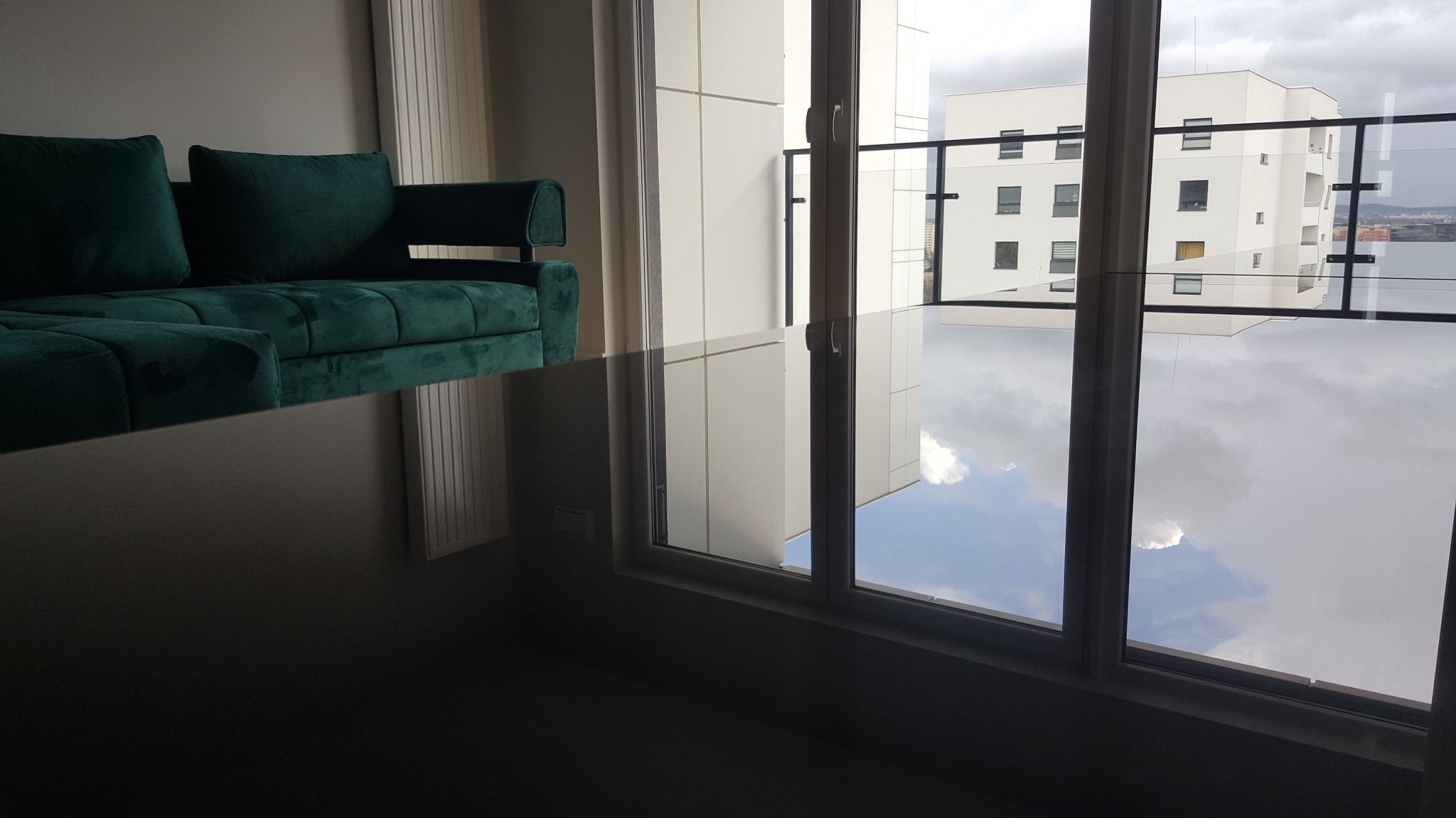 Szmaragdowa sofa narożna z weluru w nowoczesnym apartamencie, widok na budynek przez duże okno z odbiciem chmur.