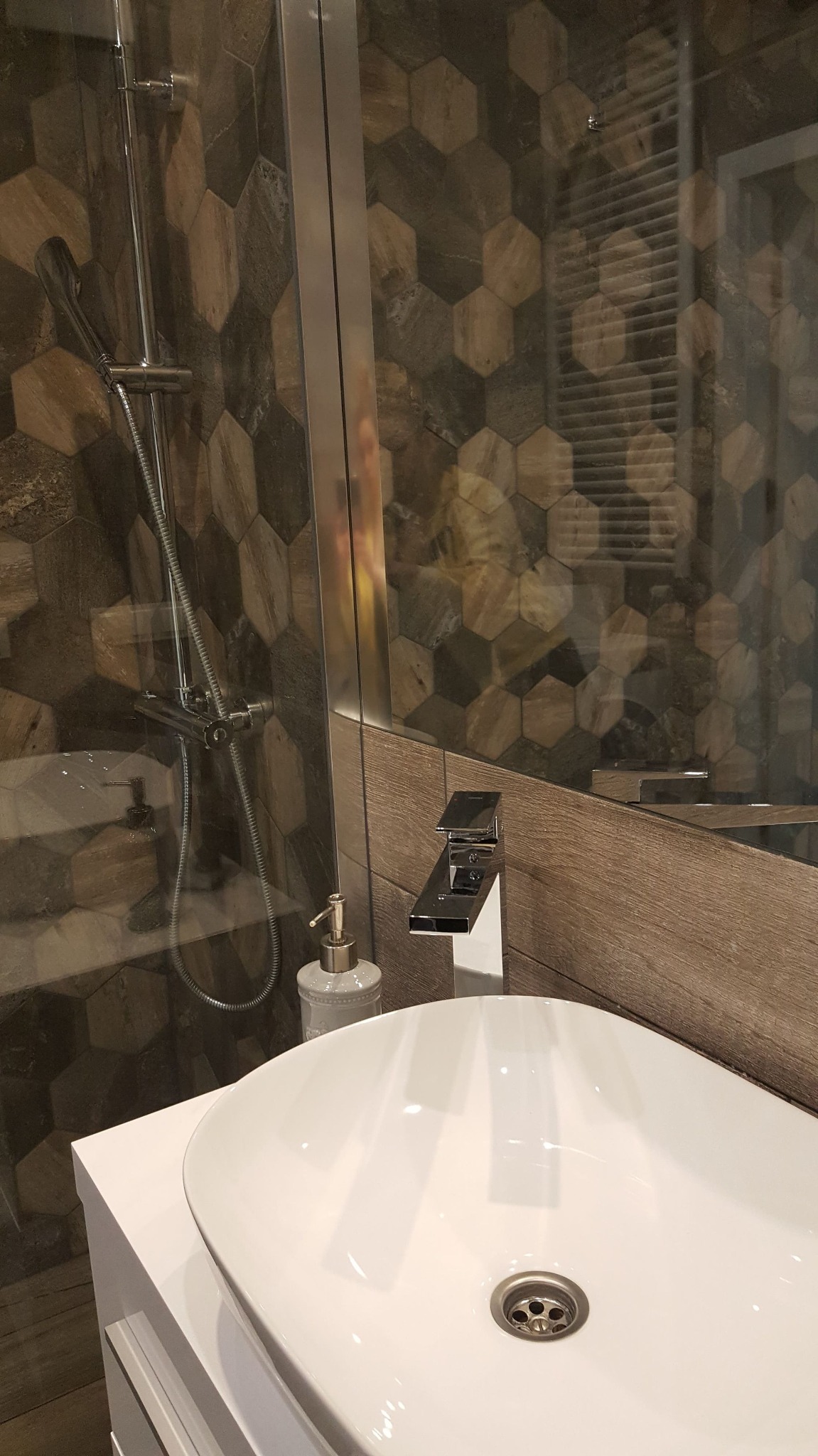 Nowoczesna łazienka z umywalką nablatową, chromowaną baterią i szklaną kabiną prysznicową z heksagonalnymi płytkami w odcieniach brązu i szarości.