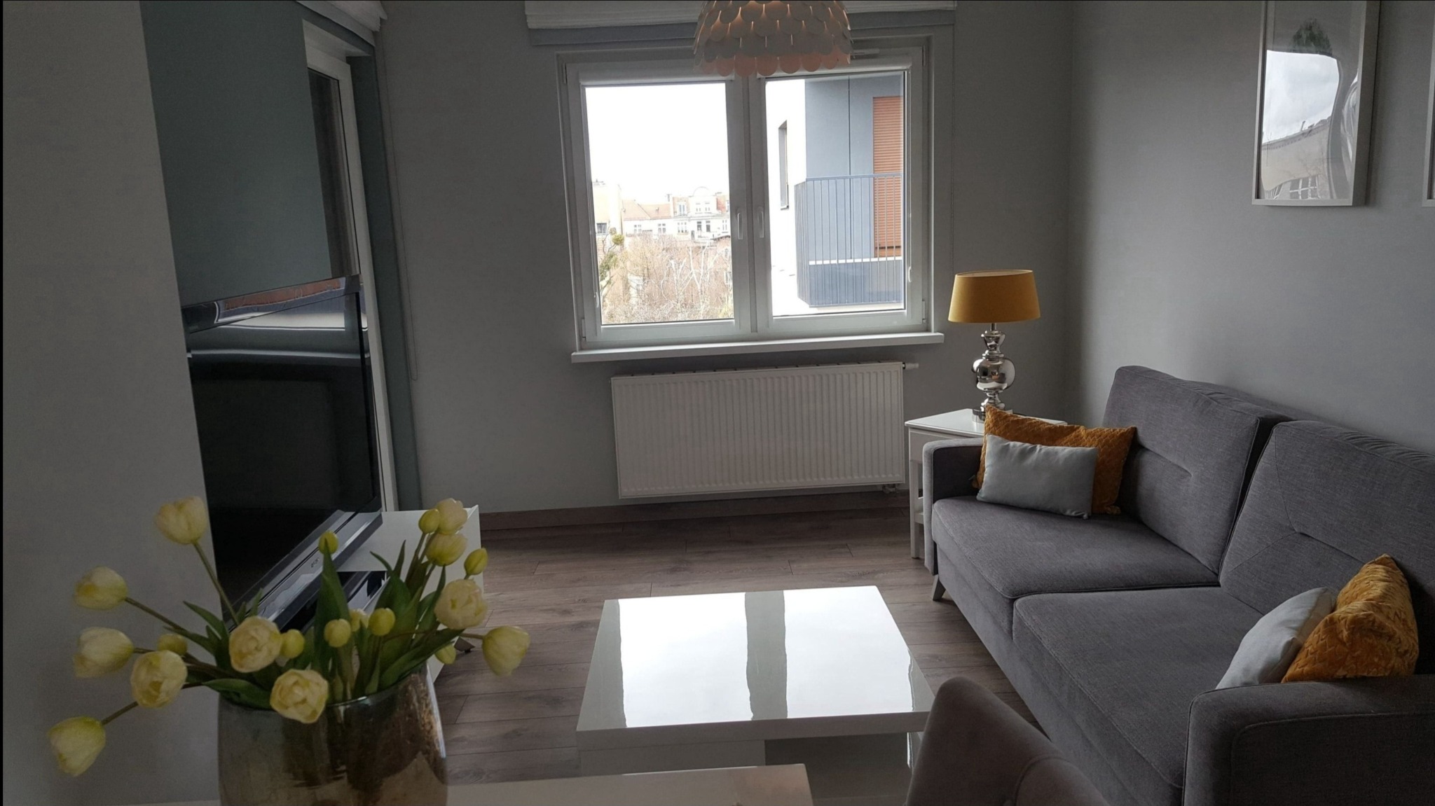 Jasne wnętrze salonu w odcieniach szarości z szarą sofą, białym stolikiem kawowym z połyskiem, bukietem kremowych tulipanów i widokiem z okna na budynki oraz drzewa bez liści.