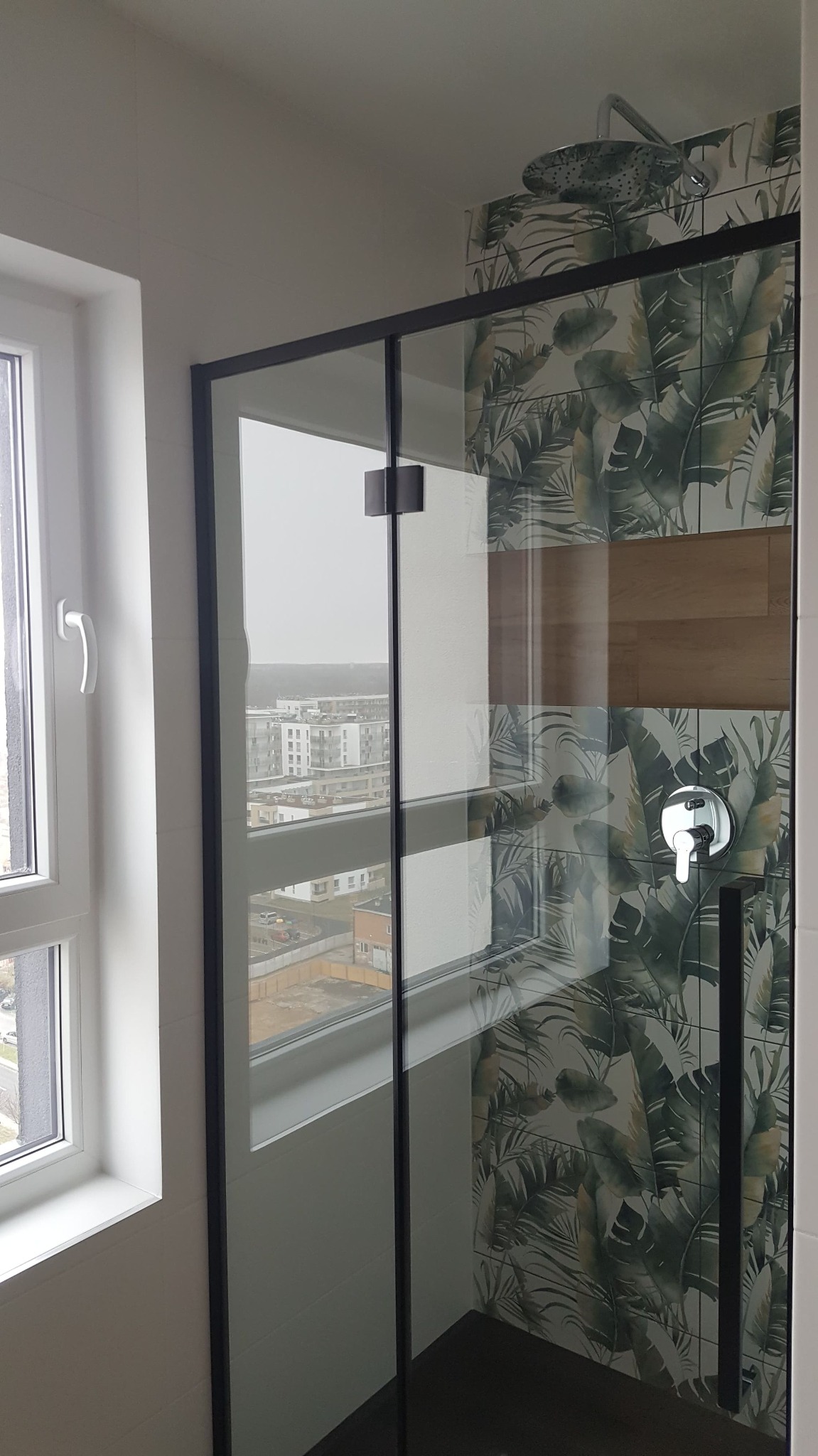 Nowoczesna kabina prysznicowa z czarnymi ramami i motywem tropikalnych liści na ścianie, widok na miasto przez okno.