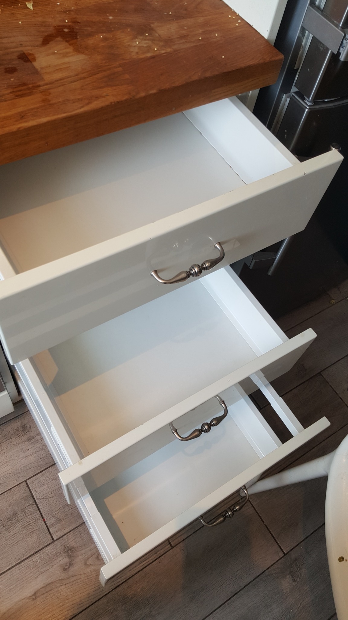 Otwarty biały regał z trzema szufladami z ozdobnymi metalowymi uchwytami na tle drewnianej podłogi i blatu.