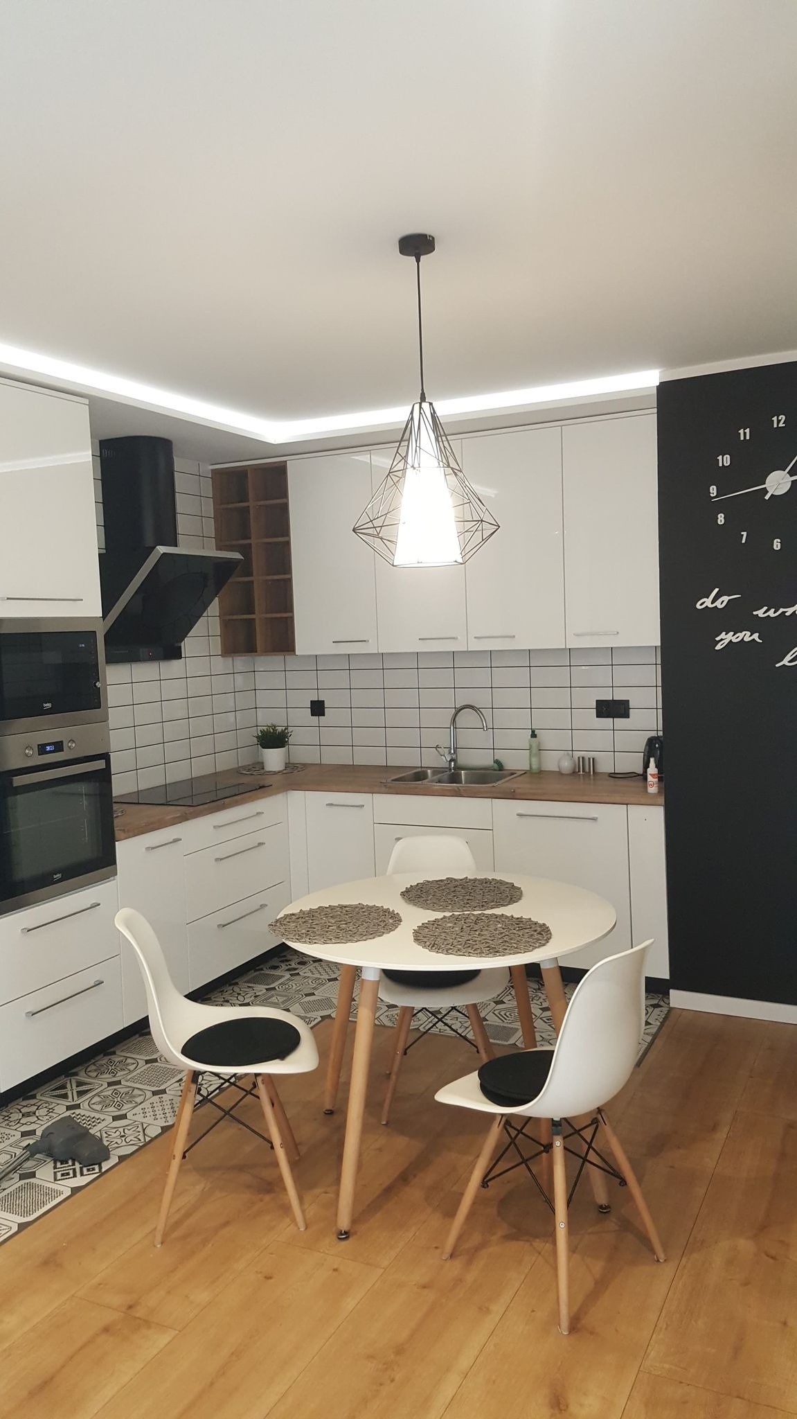 Jasna kuchnia z białymi meblami, okrągłym stołem z trzema plecionymi podkładkami i trzema krzesłami z czarnymi poduszkami, czarny zegar ścienny z napisem.