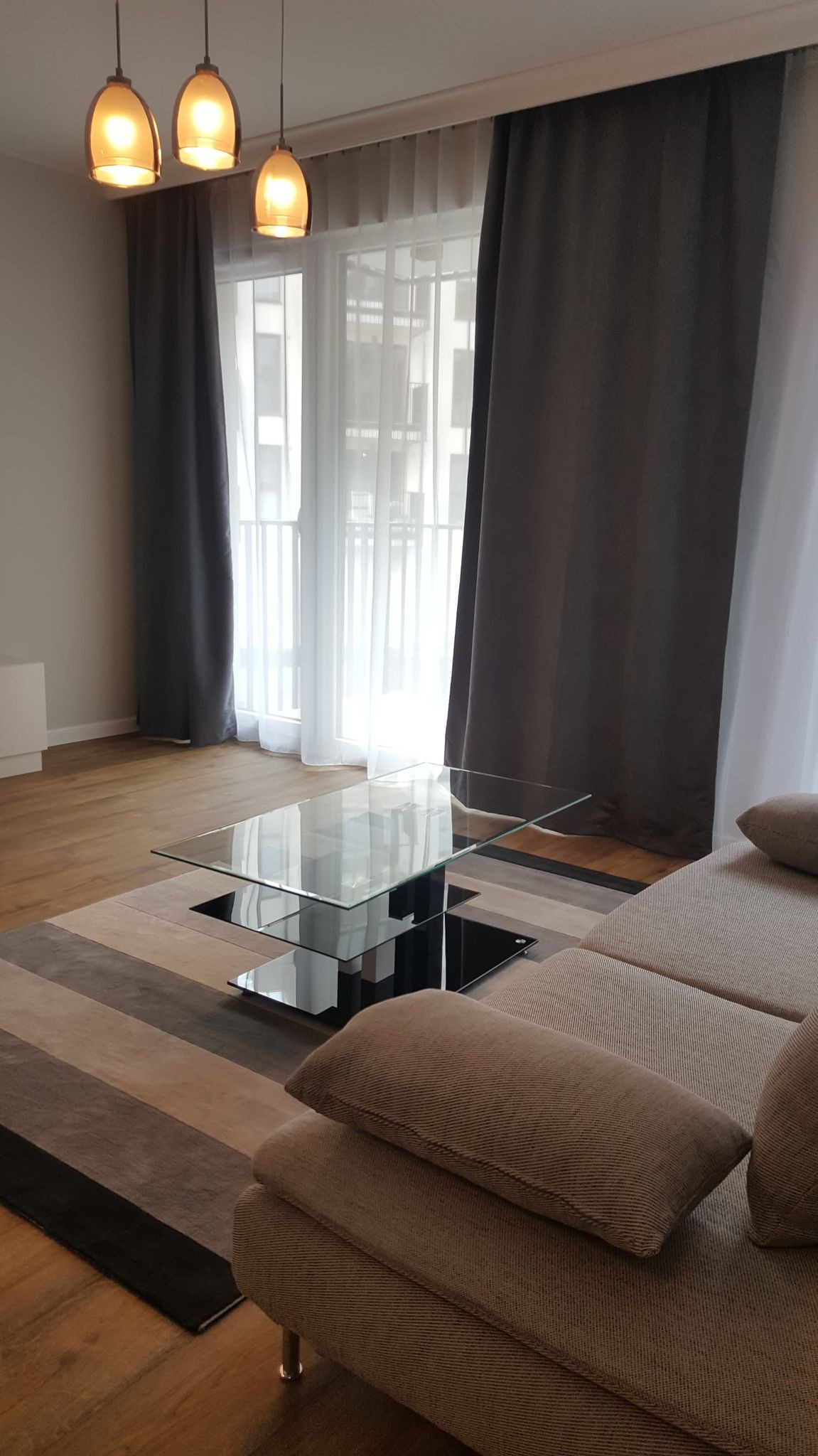 Salon z beżową sofą, szklanym stolikiem kawowym na designerskim dywanie w pasy, szare zasłony i wiszące lampy z ciepłym światłem.